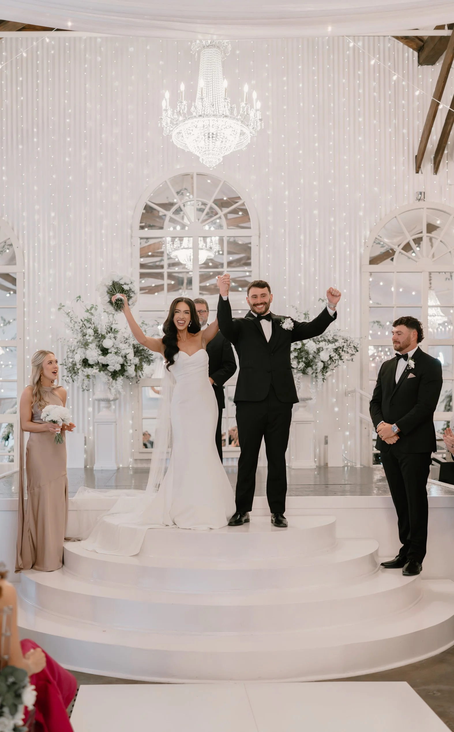 A newlywed couple celebrating on a wedding ceremony stage with raised arms. The bride in a white wedding gown and the groom in a black tuxedo. Family and friends are nearby, and the setting is decorated with white flowers, fairy lights, and chandelie