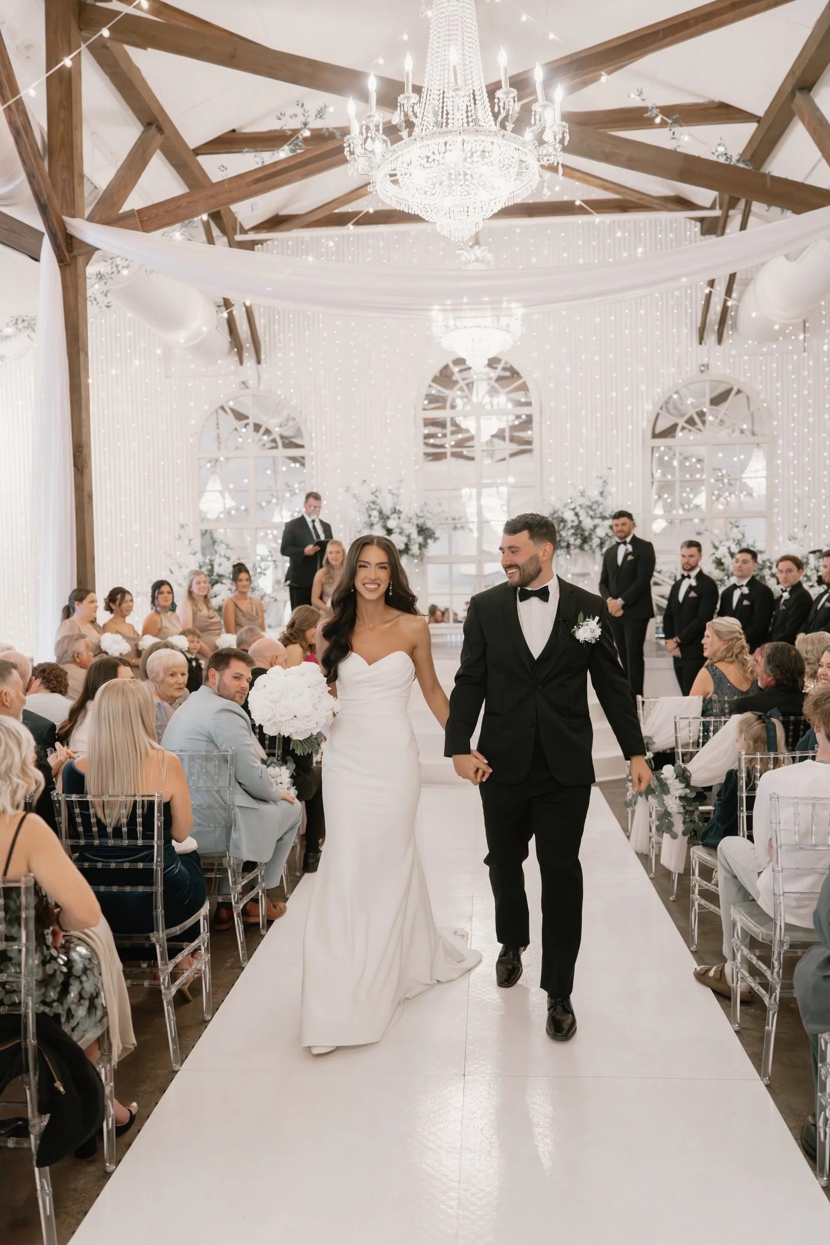 A bride and groom walking down the aisle at their wedding ceremony at Supposey warehouse, smiles on their faces, surrounded by seated guests and an elegant, decorated wedding venue with a chandelier and string lights.