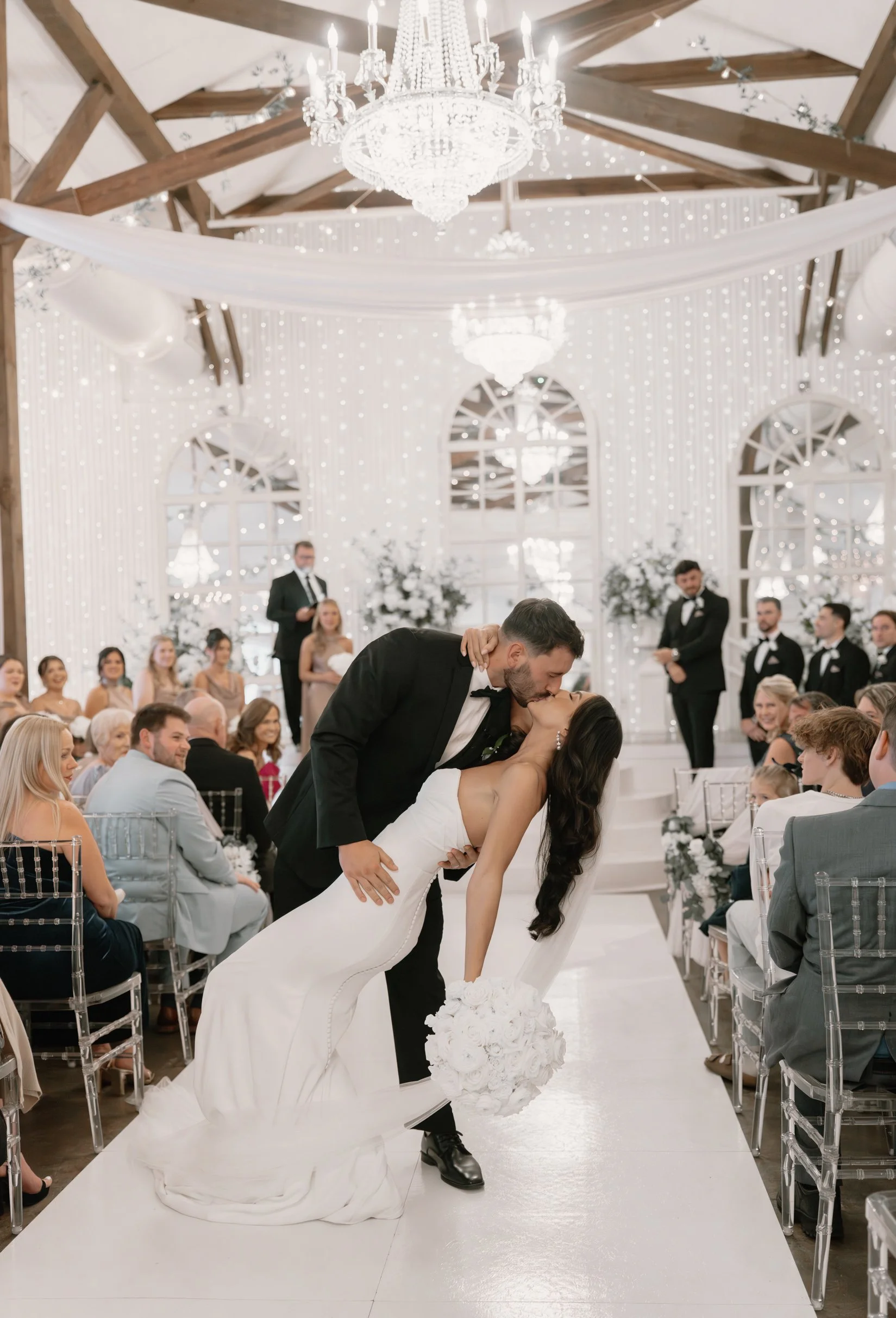 A bride and groom kiss during their wedding ceremony in an elegant, white decorated Pensacola venue with chandeliers and string lights, surrounded by guests seated on transparent chairs.