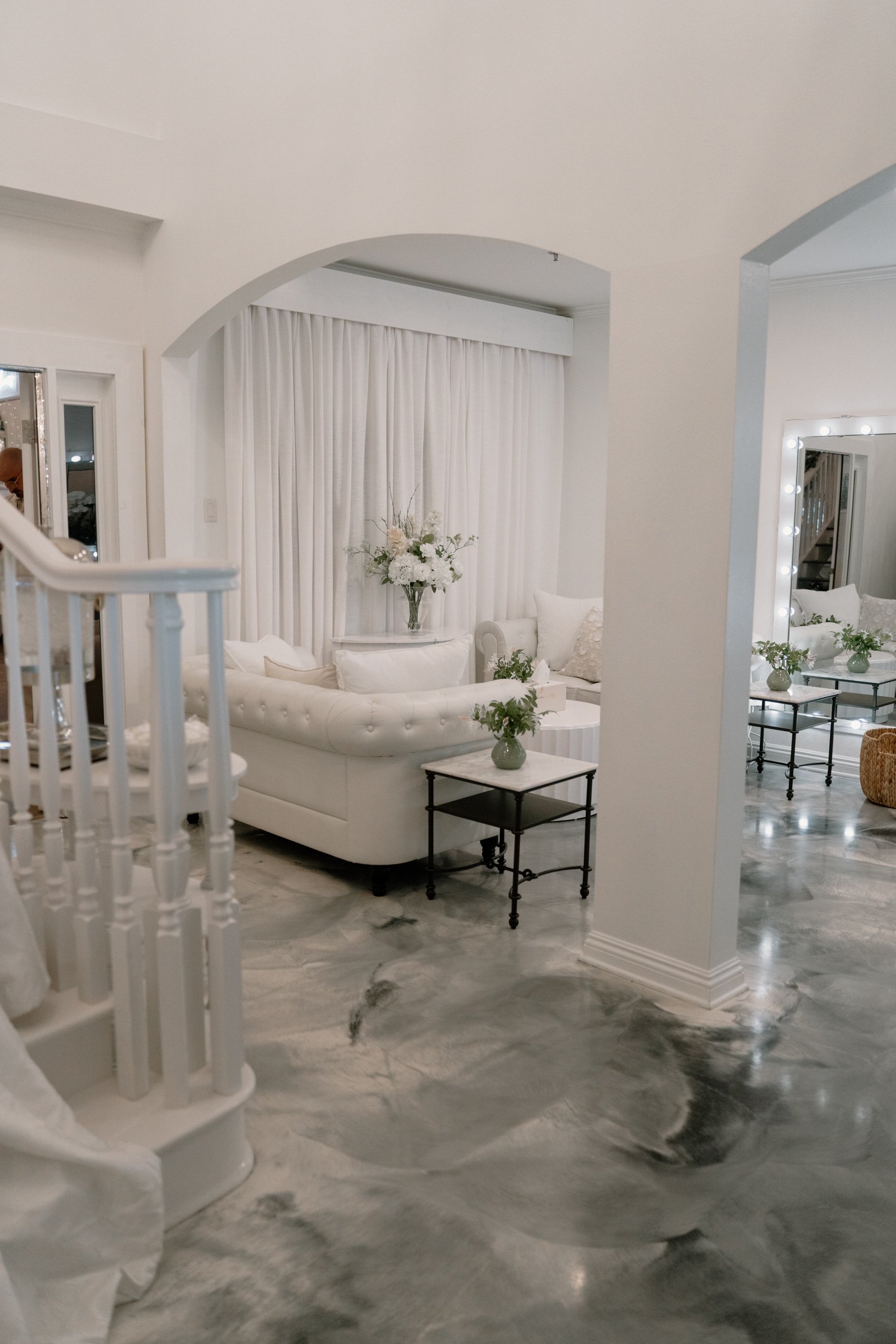 bridal suite with white tufted sofa, marble floor, and white curtains. Decor includes vases with flowers, and a mirror with lights.
