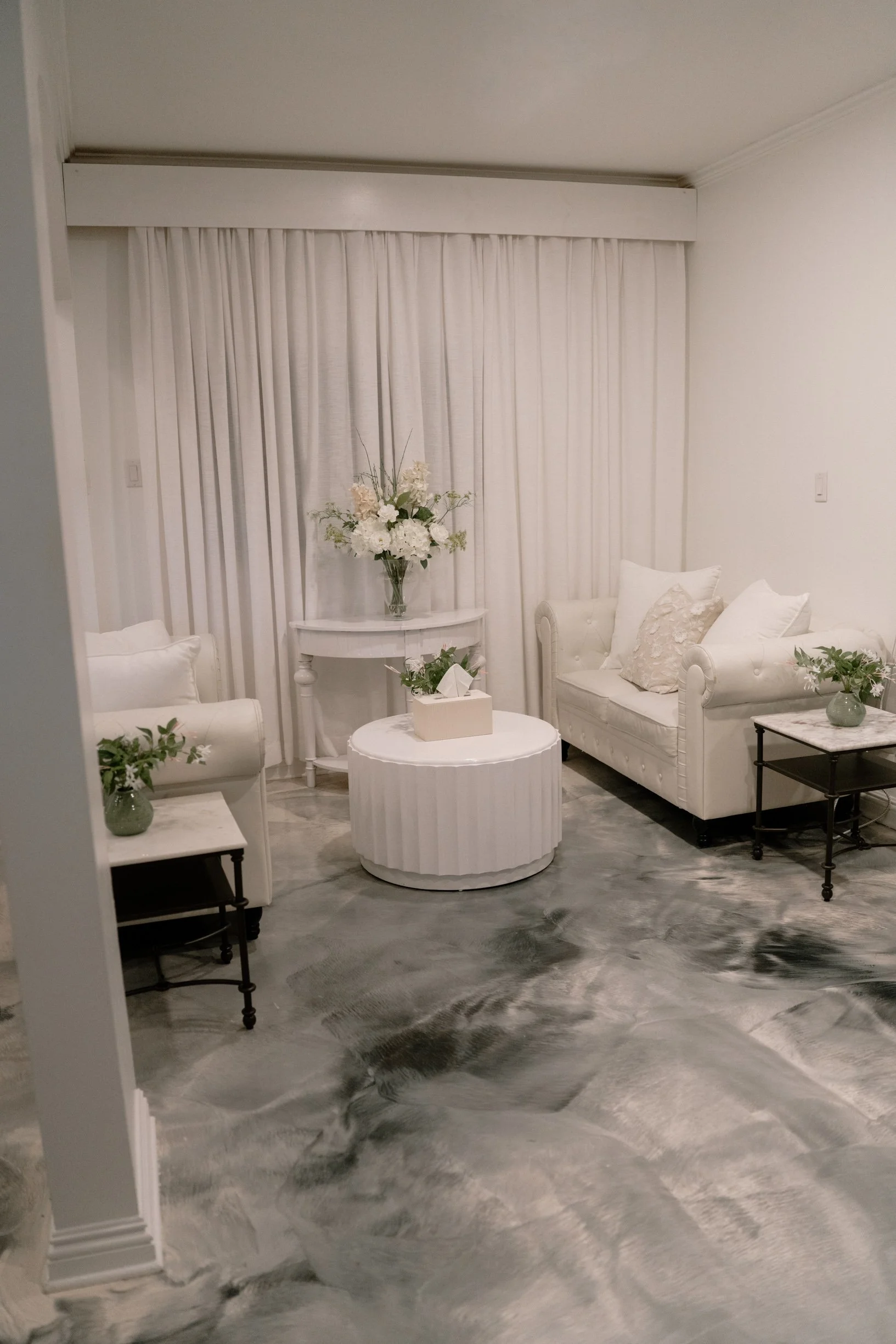A cozy, elegant bridal suite decorated in white with floral arrangements, white curtains, two white sofas with pillows, and a marbled floor.