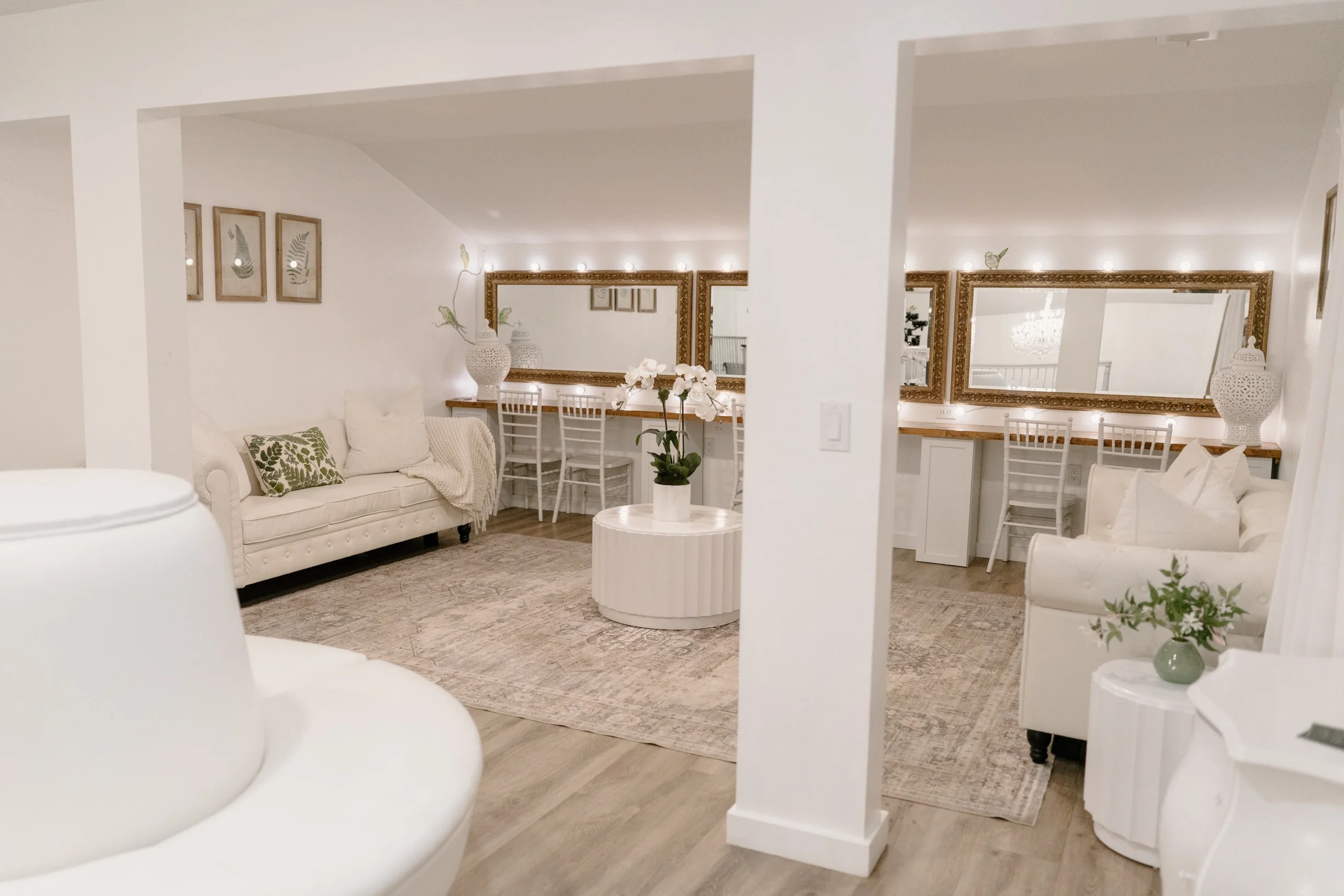 A bright, elegant bridal suite with white sofas, a round coffee table with an orchid, large mirrors with gold frames, and a built-in desk area with white chairs and decorative vases.