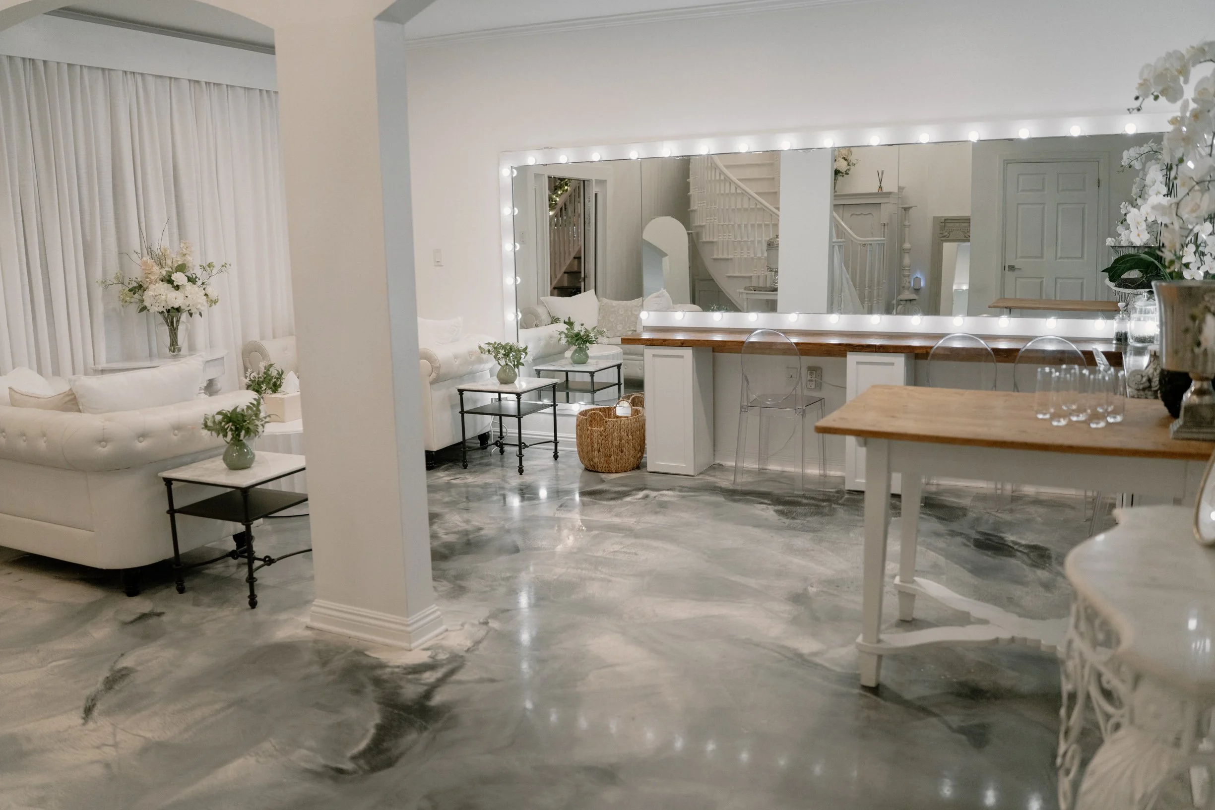 A bridal suite with white and neutral decor, large mirror with light bulbs, marble floor, white sofas, glass tables, and floral arrangements.