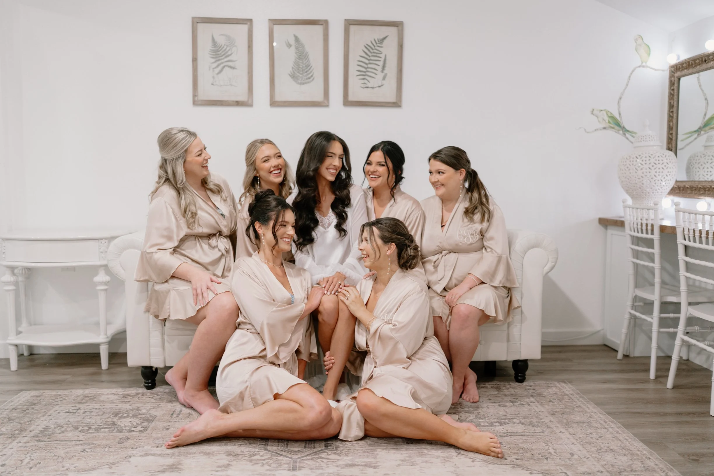 A bride and her six bridesmaids on a white sofa, all wearing matching satin robes, smiling and laughing during a bridal party gathering in a decoratively styled bridal suite.