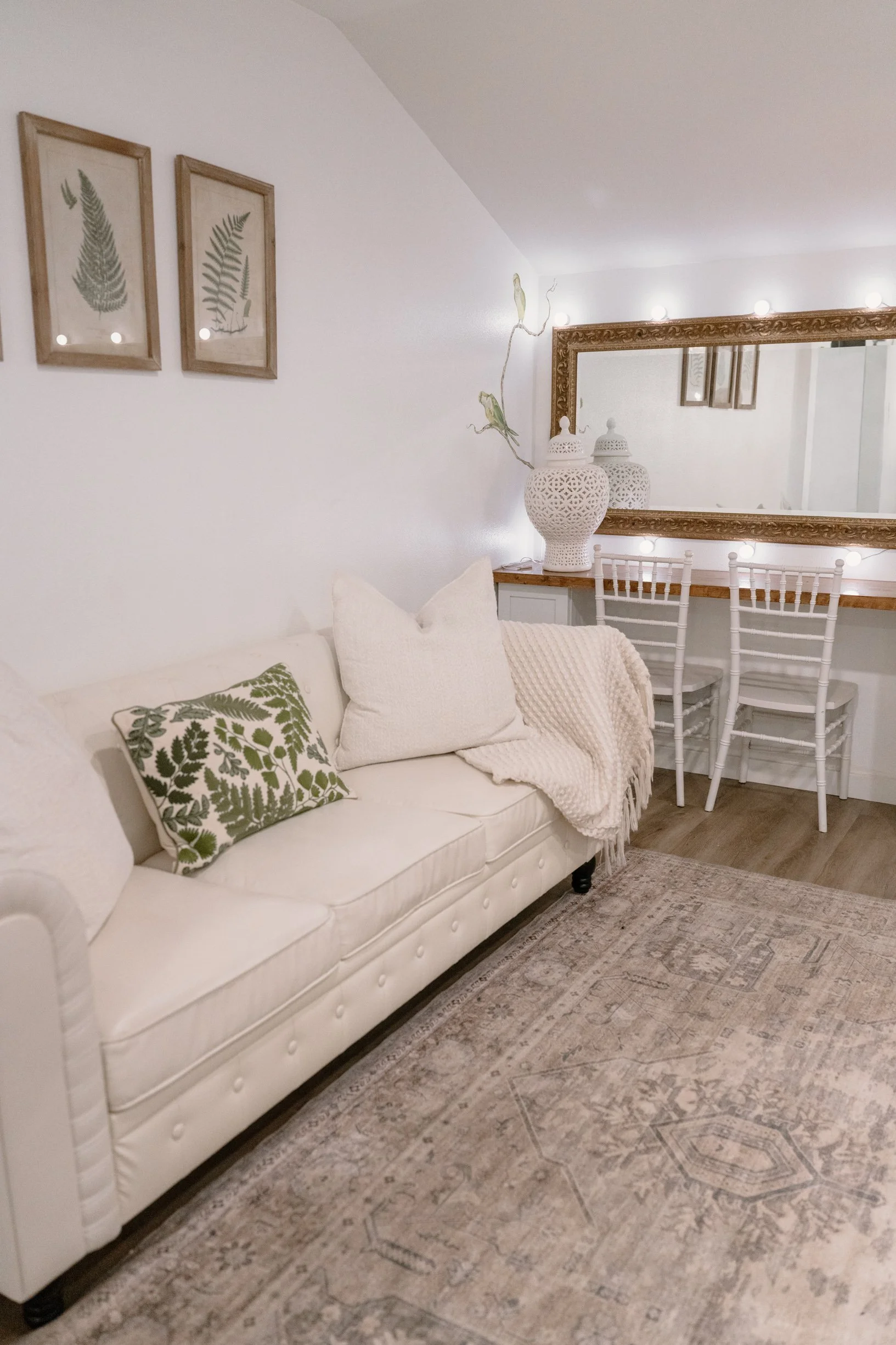 bridal suite corner with white sofa, green fern print pillows, beige throw blanket, floral wall art, large ornate mirror, white decorative vases, and chairs in the background.