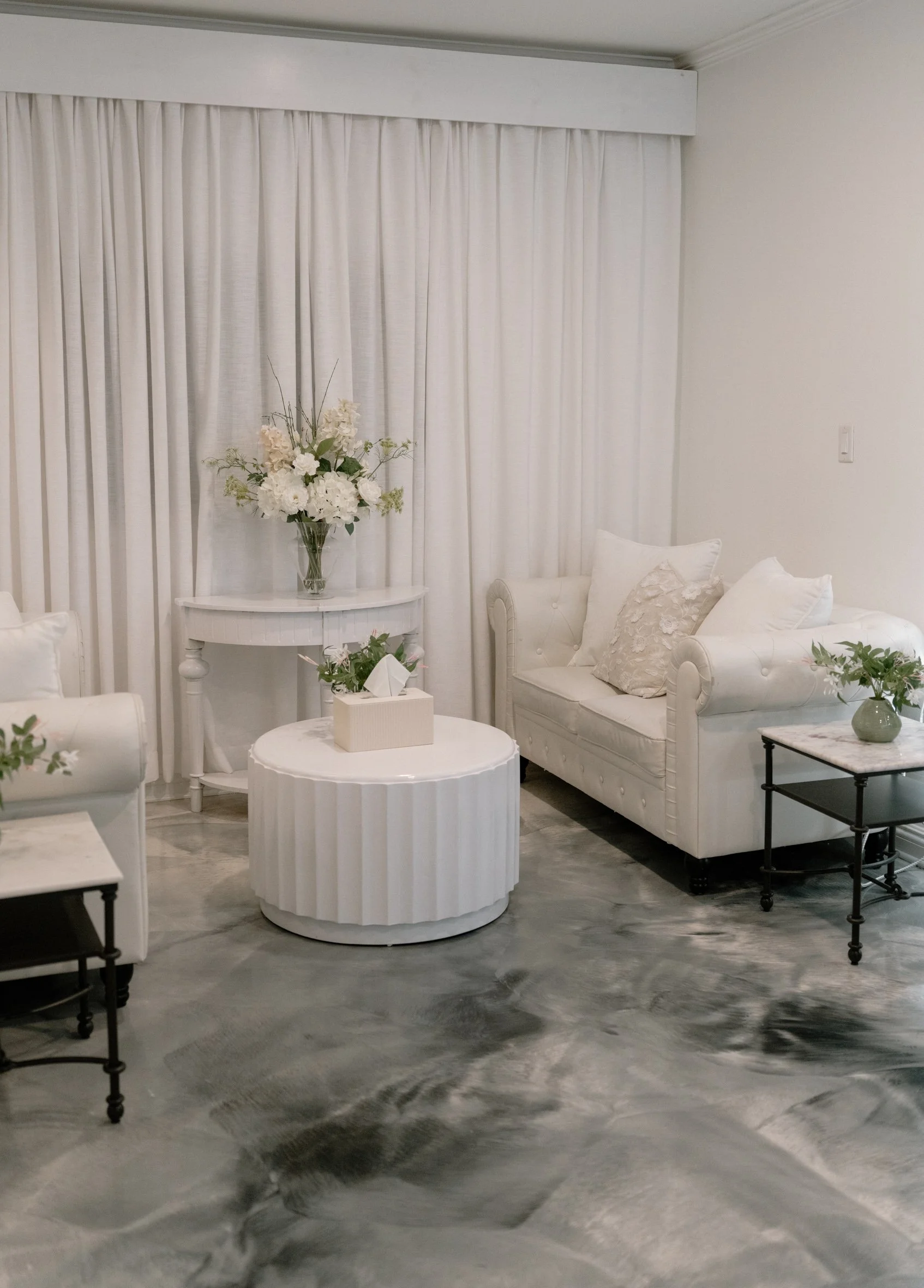 A living room with white furniture, including a tufted white sofa with decorative pillows, a white side table with a green vase, a white round coffee table with a tissue box, and a white console table with a large flower arrangement in a glass vase. 