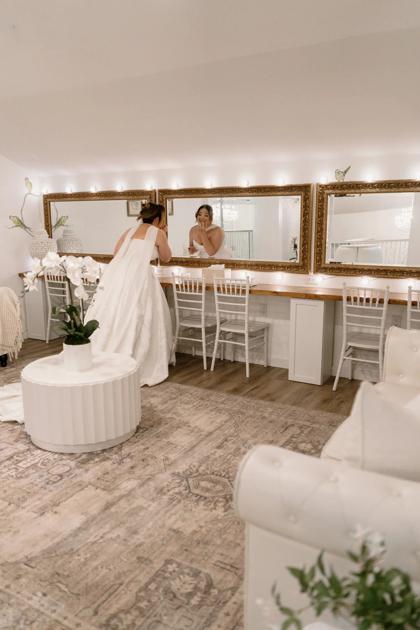 A bride in a white wedding dress standing in front of a large mirror, smiling and holding her hand near her face, in a well-lit bridal suite decorated with white flowers and elegant decor.