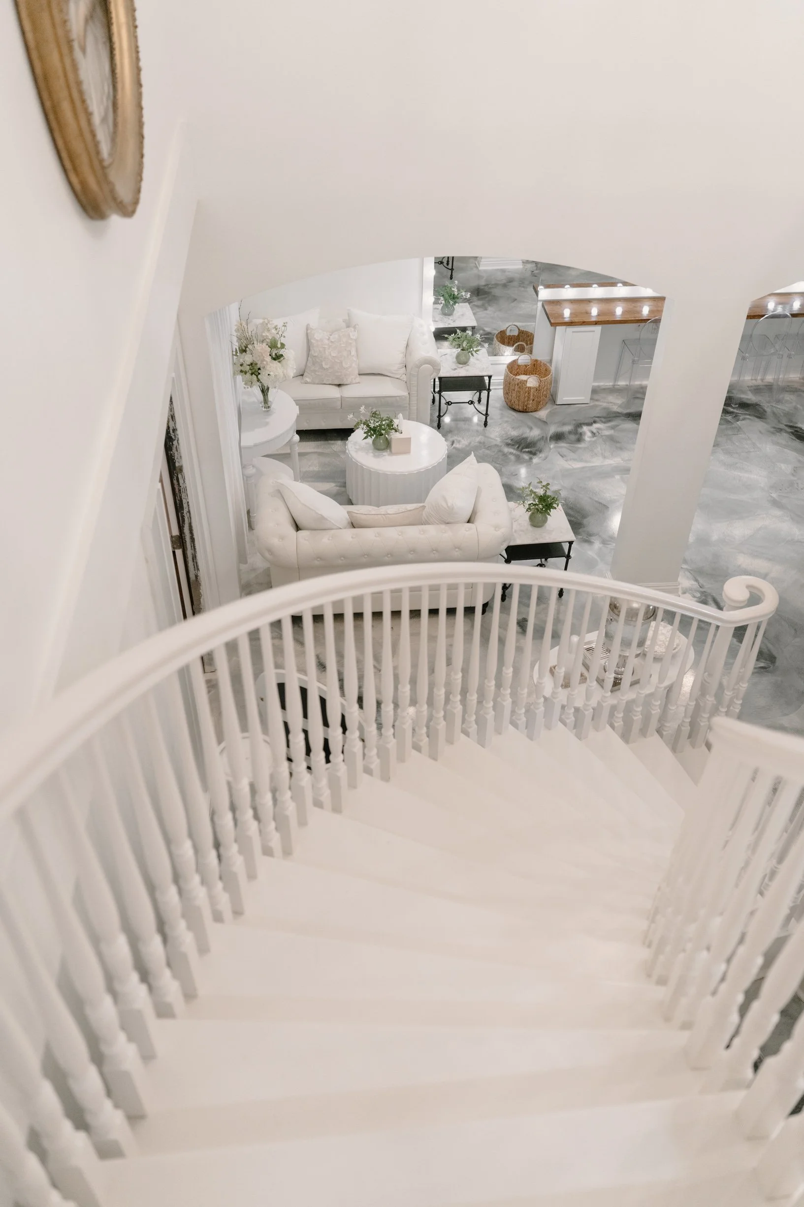 Looking down from the top of a white staircase into a modern bridal suite with white furniture, plants, and a marble floor.
