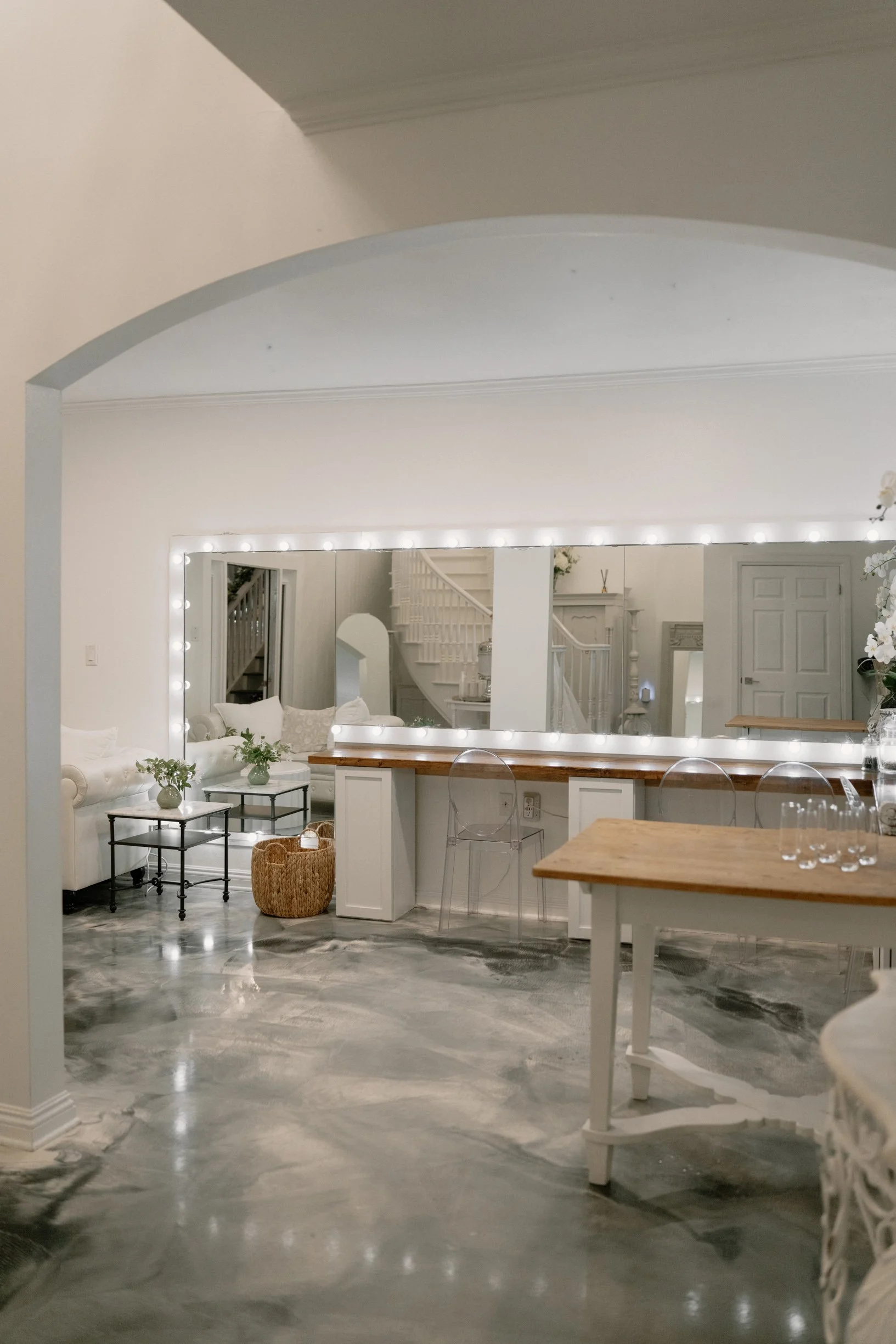 A spacious, modern bridal suite with a large mirror framed with lights, a white sofa, small tables with green plants, a woven basket, a marble floor, and a wooden bar with transparent chairs.
