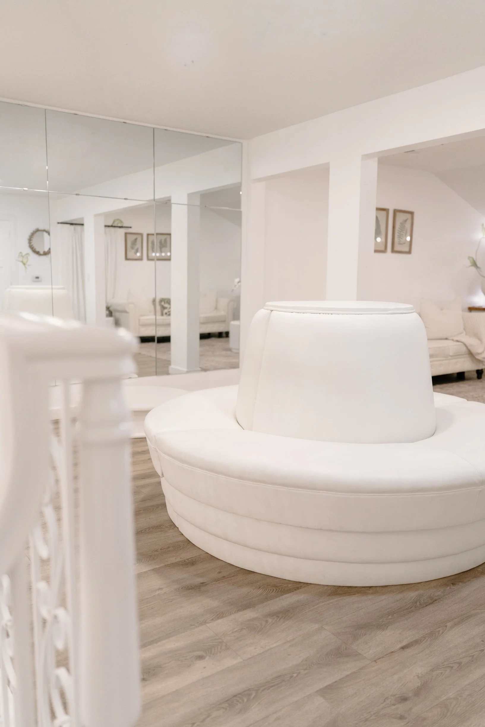 A white circular chair with a high back, positioned in a bridal suite with beige wood flooring, mirrored closet doors, white walls, and contemporary wall art.