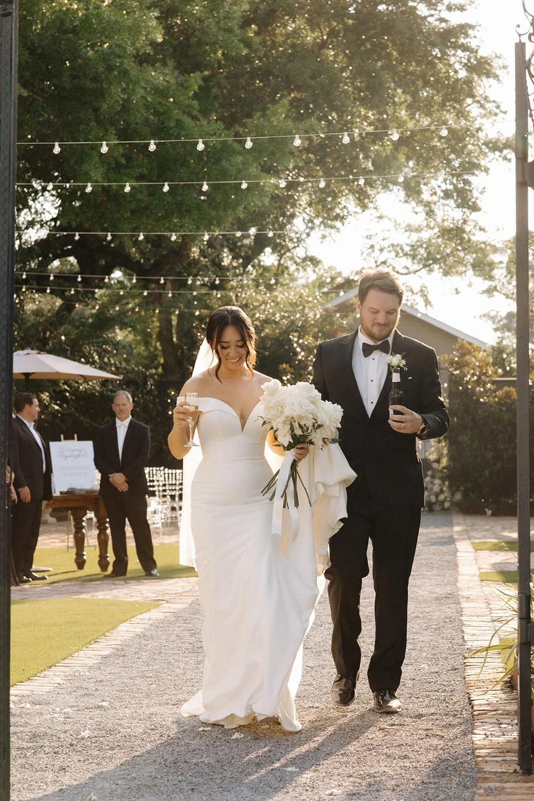 Bride and groom walking together outdoors during Pensacola wedding celebration, the bride holding a bouquet and both holding drinks, with guests in the background.