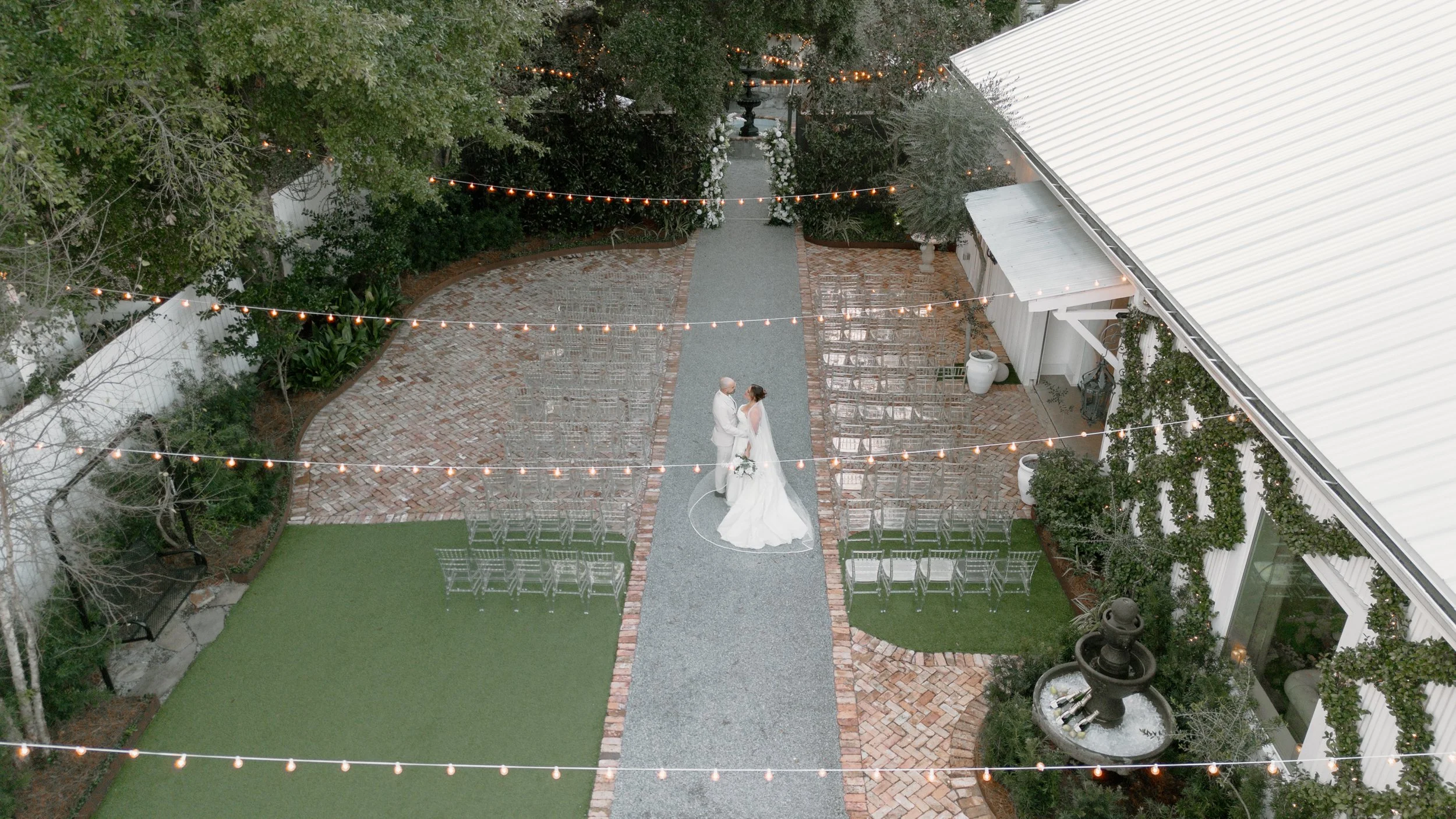 A bride and groom stand together in the center of an outdoor Pensacola wedding ceremony area, surrounded by string lights, chairs, and lush greenery.
