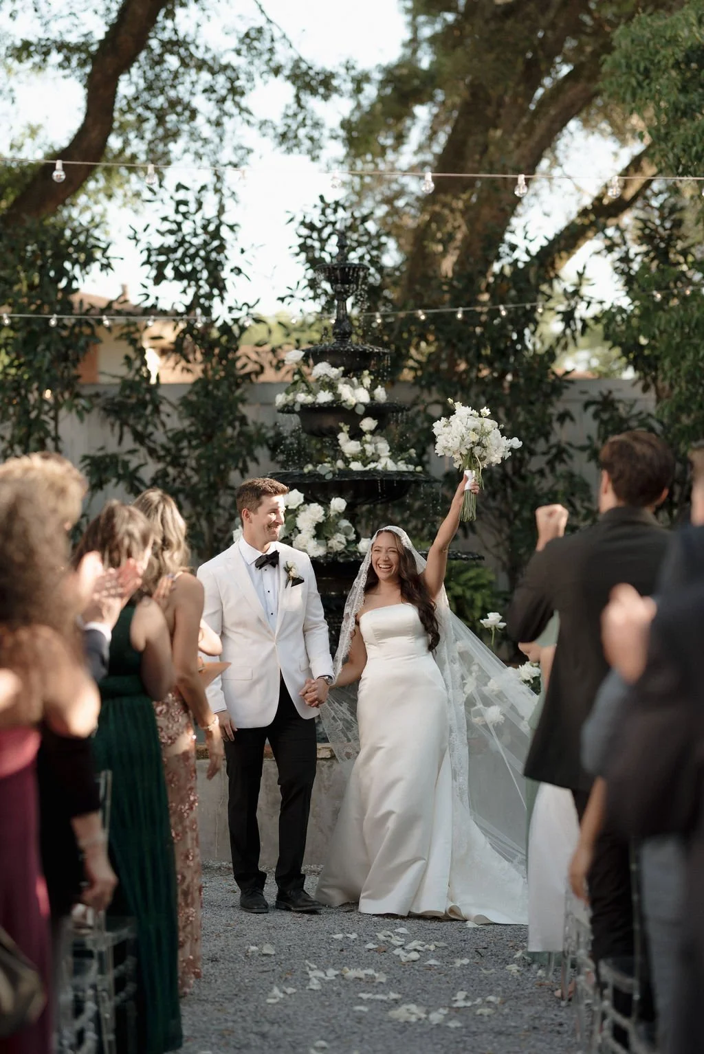 A bride and groom hold hands at their outdoor Pensacola wedding ceremony, with the bride raising a bouquet in celebration, surrounded by friends and family.