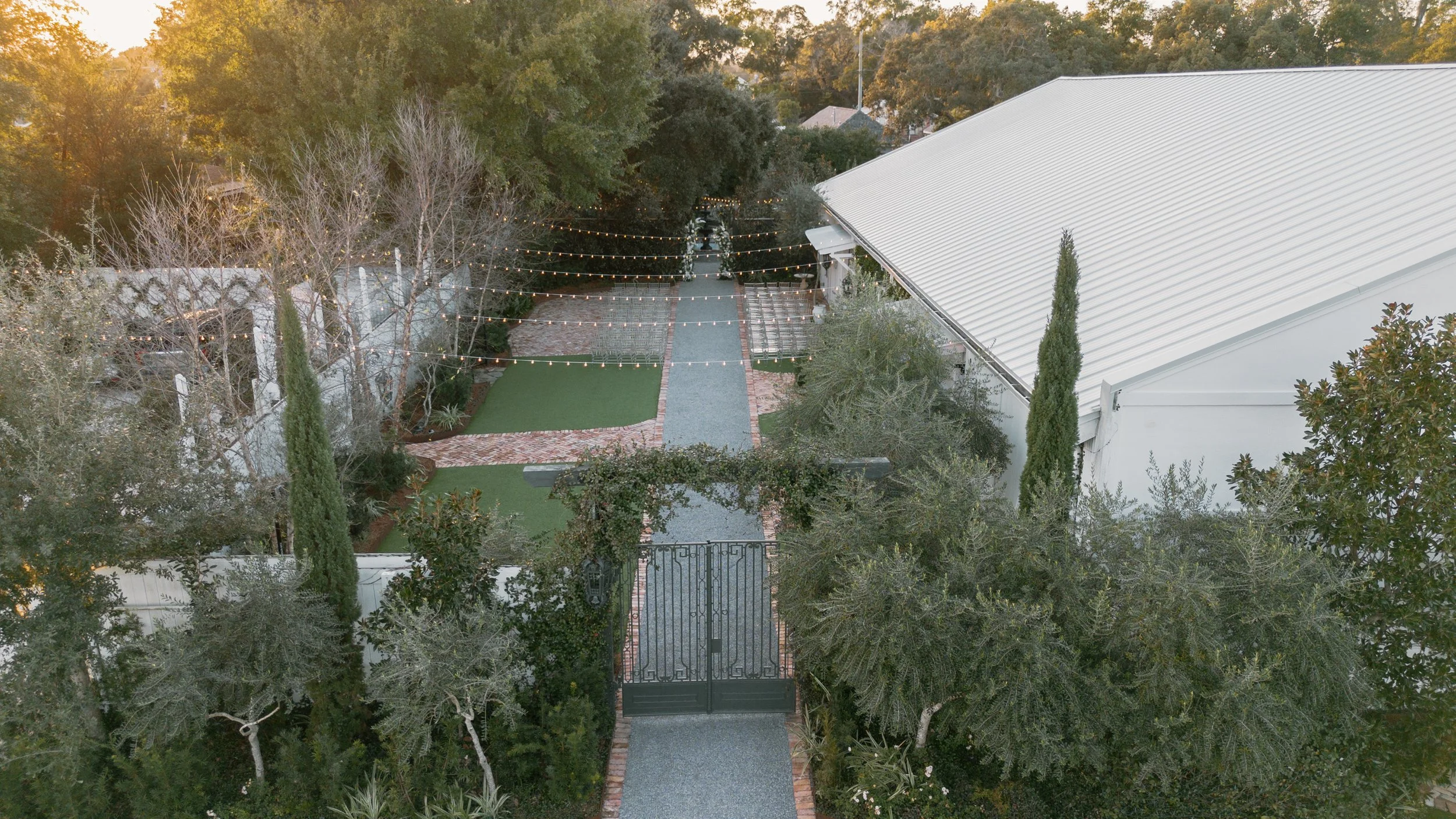 Aerial view of a Pensacola Wedding Venue garden with a pathway, string lights, trees, and a white house with a metal roof.