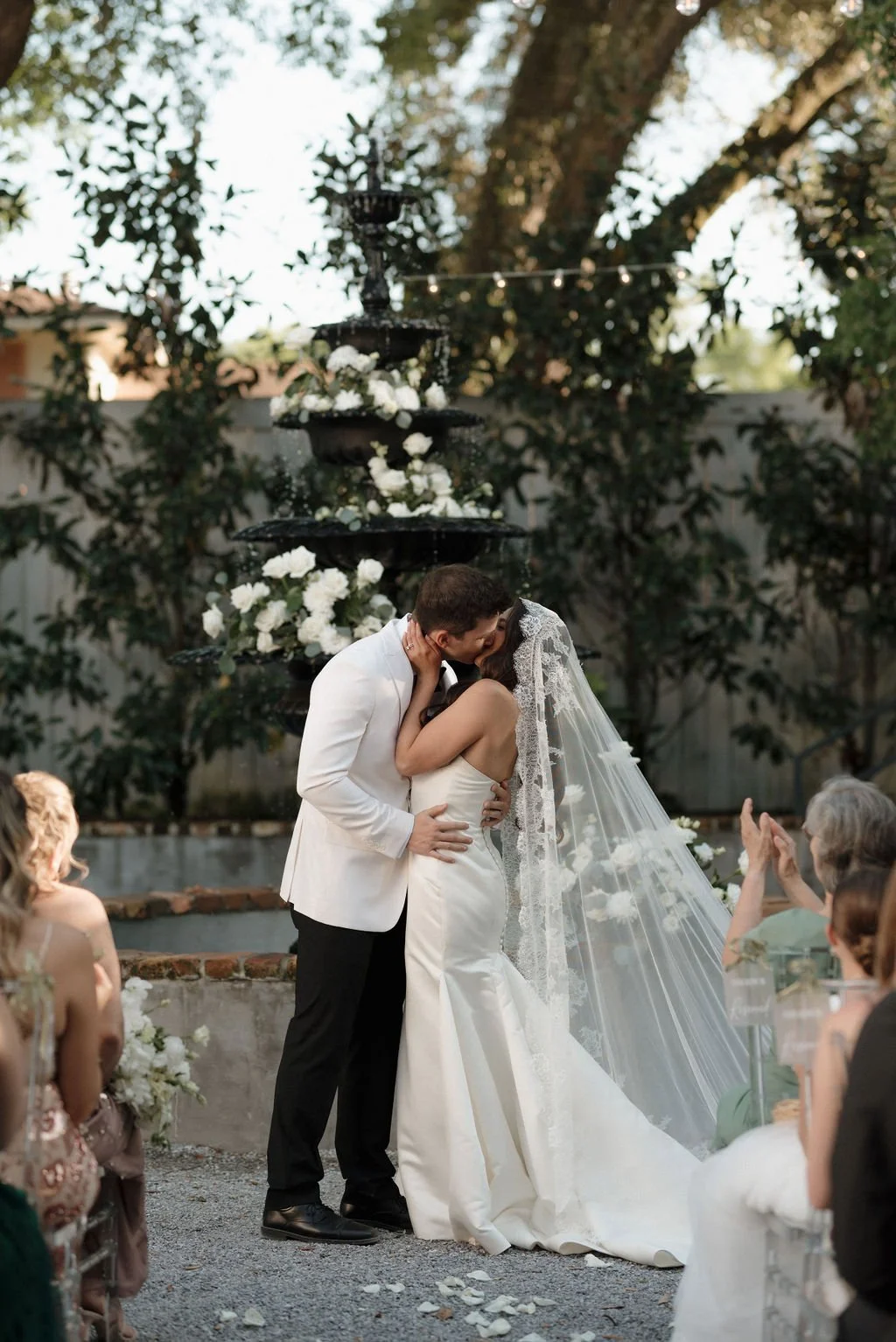A bride and groom kiss during their outdoor Pensacola wedding ceremony, with guests seated around them and a fountain with white flowers in the background.