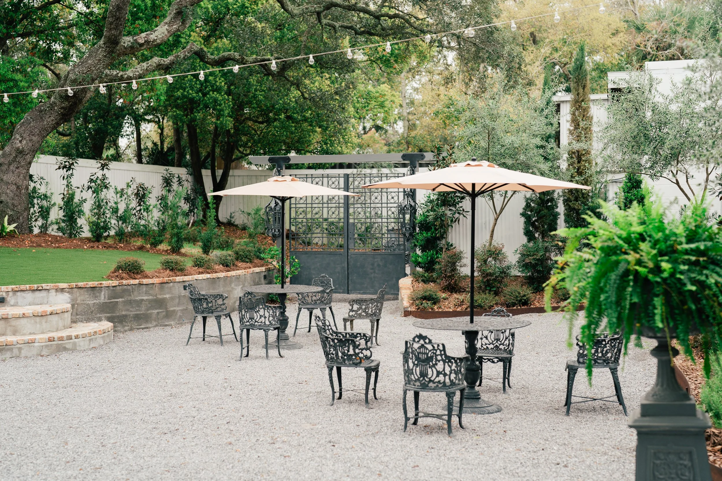 Outdoor Pensacola Wedding Venue patio area with metal table and black ornate chairs, white umbrellas, lush green trees, plants, and a gravel ground. There is a gated entrance and string lights overhead.
