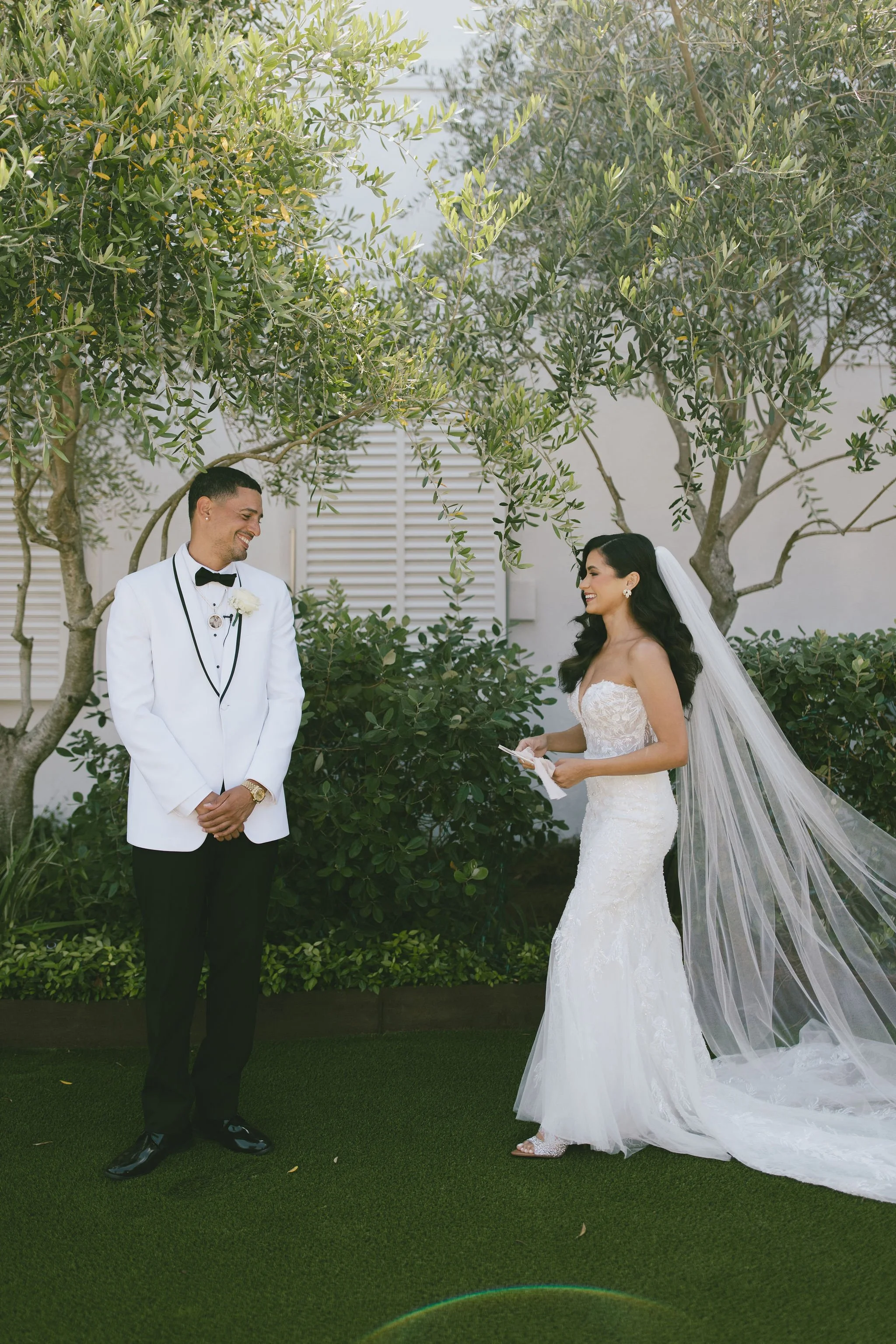A bride and groom stand outdoors under trees, exchanging vows. The groom wears a white tuxedo jacket with a black bow tie, and the bride wears a strapless lace wedding gown with a long veil. Both are smiling at each other in a Pensacola Wedding Venue