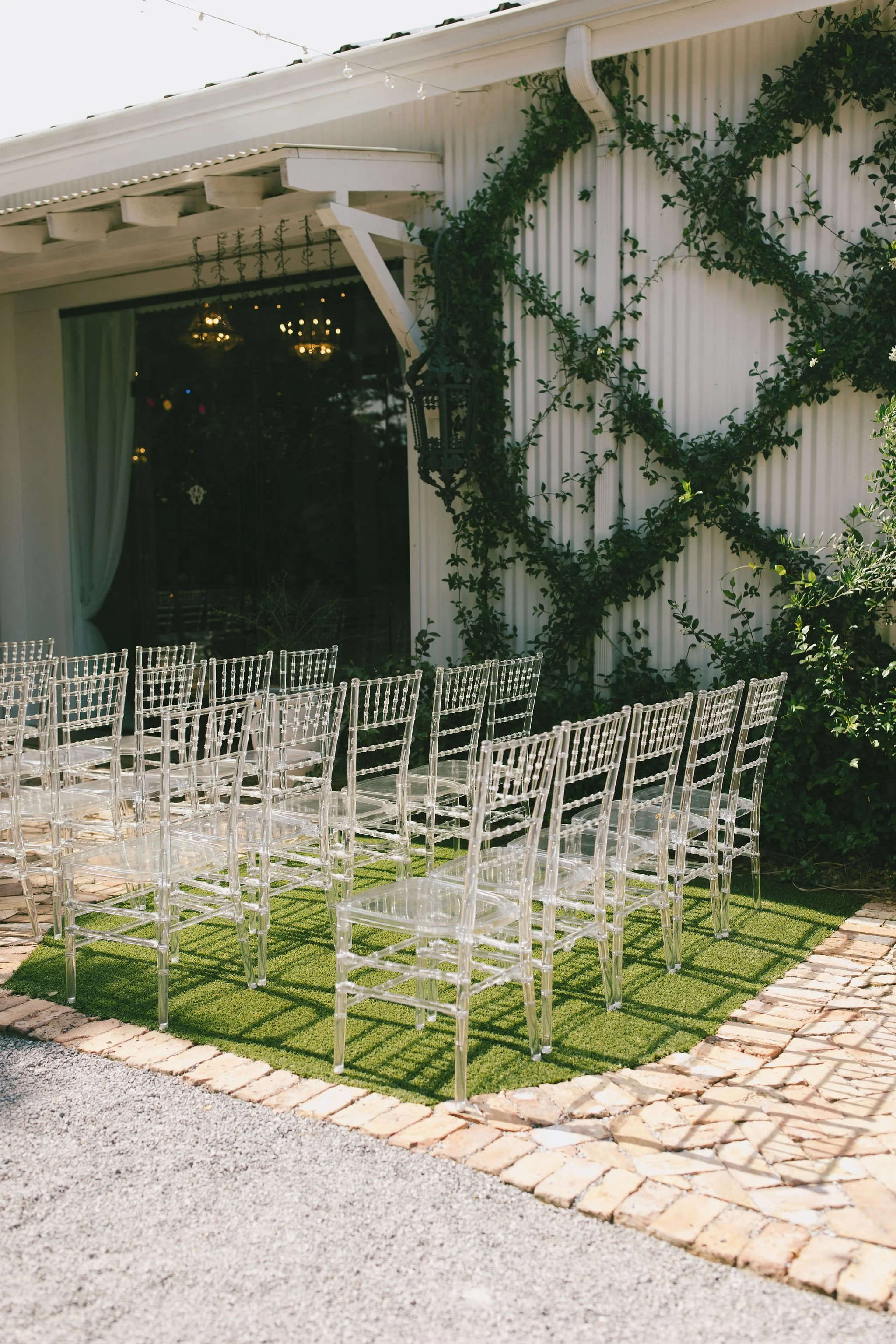 Empty clear acrylic chairs arranged outdoors on a green turf mat at a wedding or event venue, adjacent to a white building with green vines and windows with curtains in a Pensacola Wedding Venue.