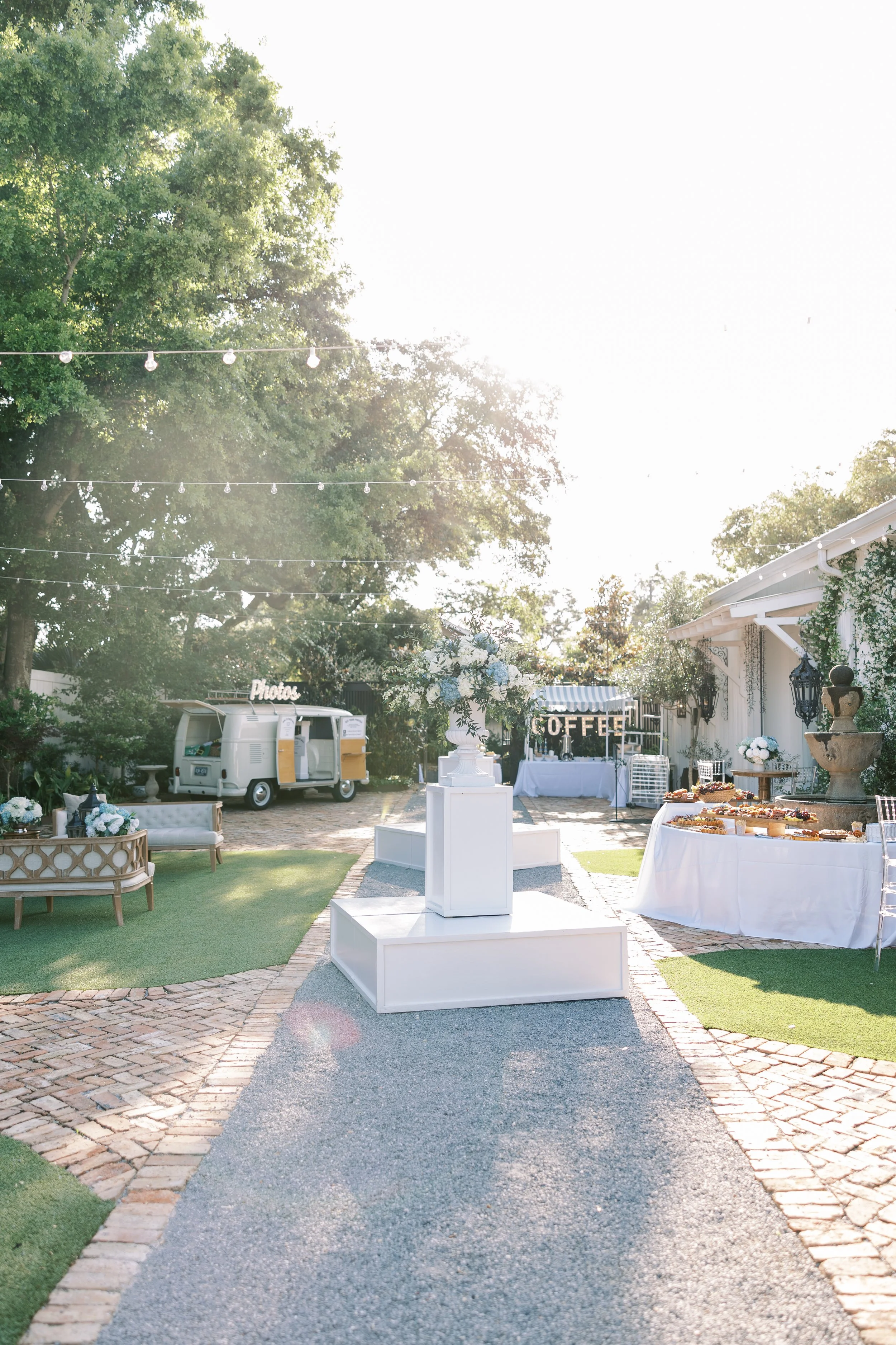 An outdoor wedding setup in a garden with string lights overhead, a white food truck, a photo booth, a tables with snacks, and a coffee station, surrounded by trees and greenery.