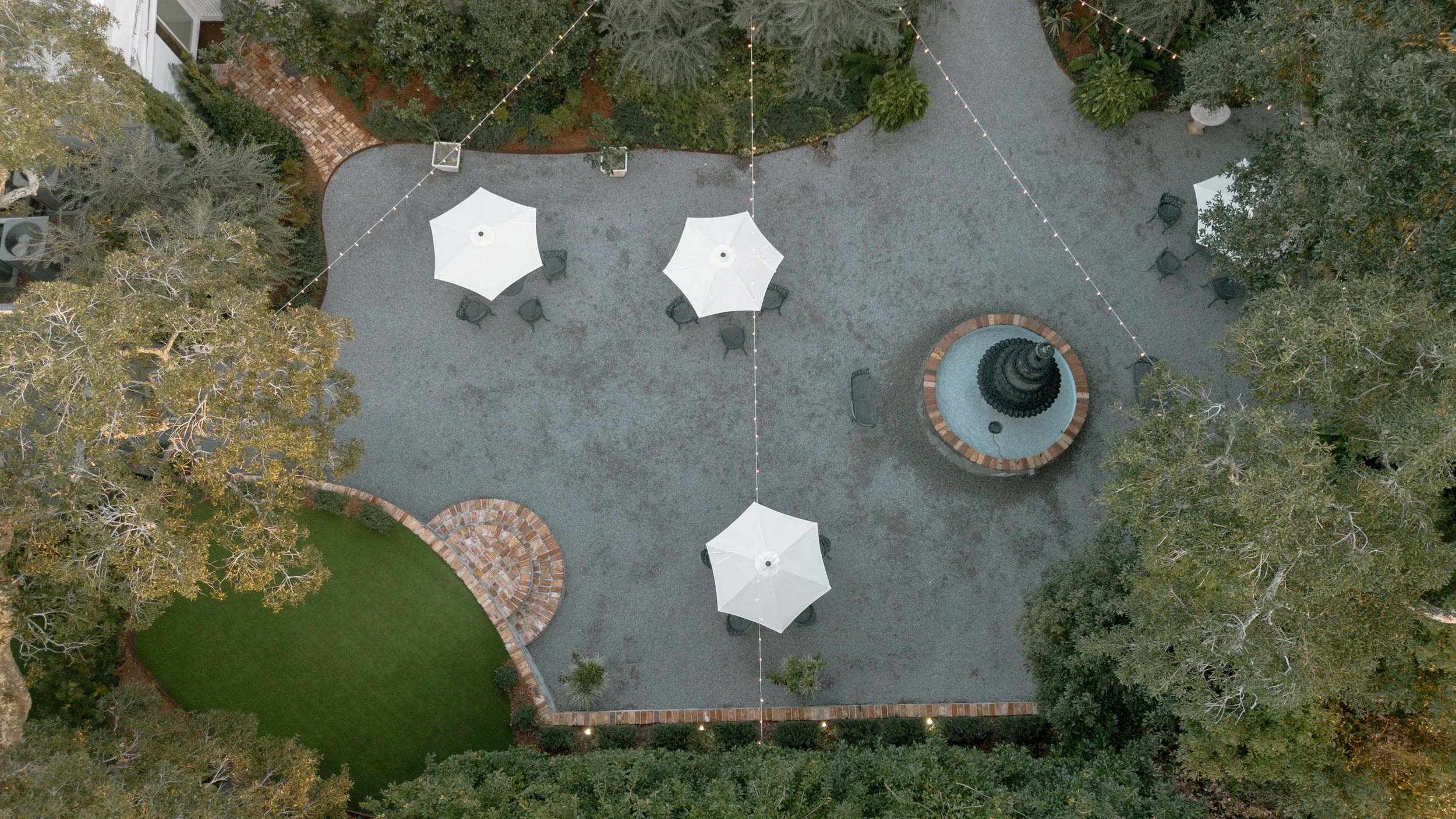 An aerial view of outdoor patio area in a Pensacola Wedding Venue with four white umbrellas, a fountain, arranged seating, string lights, and surrounding trees and landscaped areas.