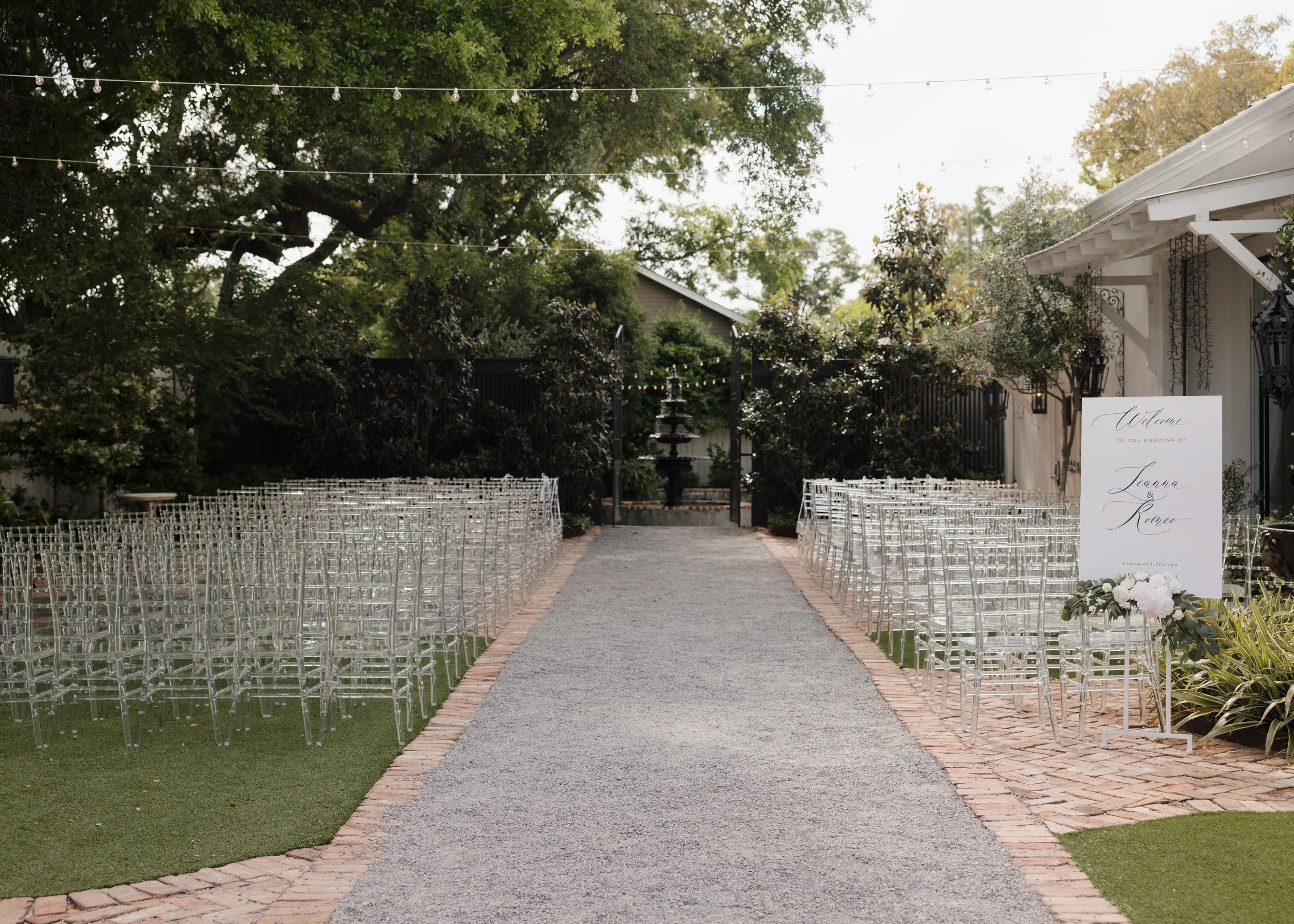 Outdoor Pensacola wedding ceremony setup with rows of transparent chairs on either side of a gravel aisle, decorative sign, lush trees, and string lights overhead.