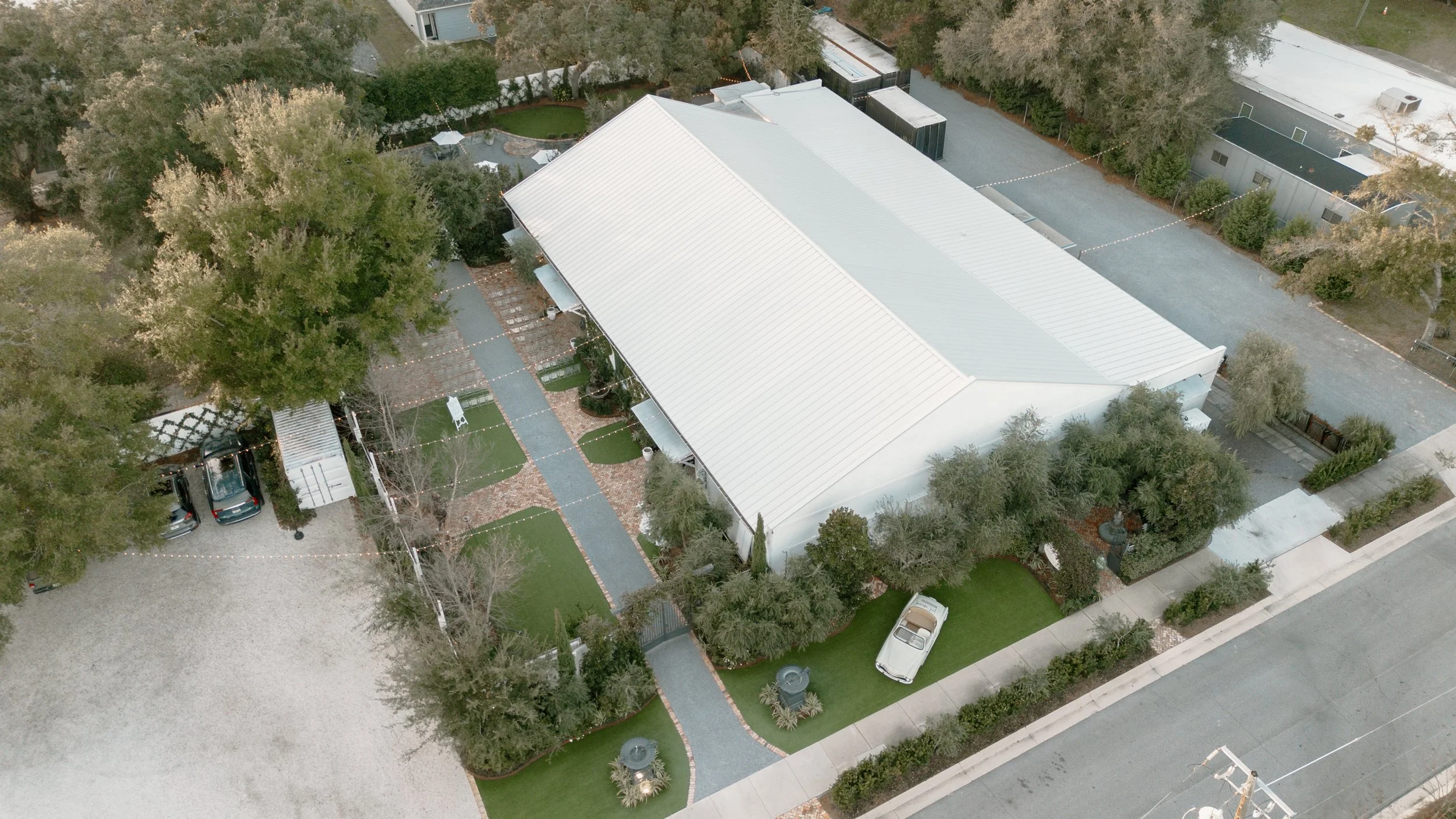 Aerial view of a large white building with a metal roof, surrounded by a garden with green grass, trees, and a paved walkway. There are parked cars, a small white carriage, and outdoor furniture visible. The building is located on a street with a sid
