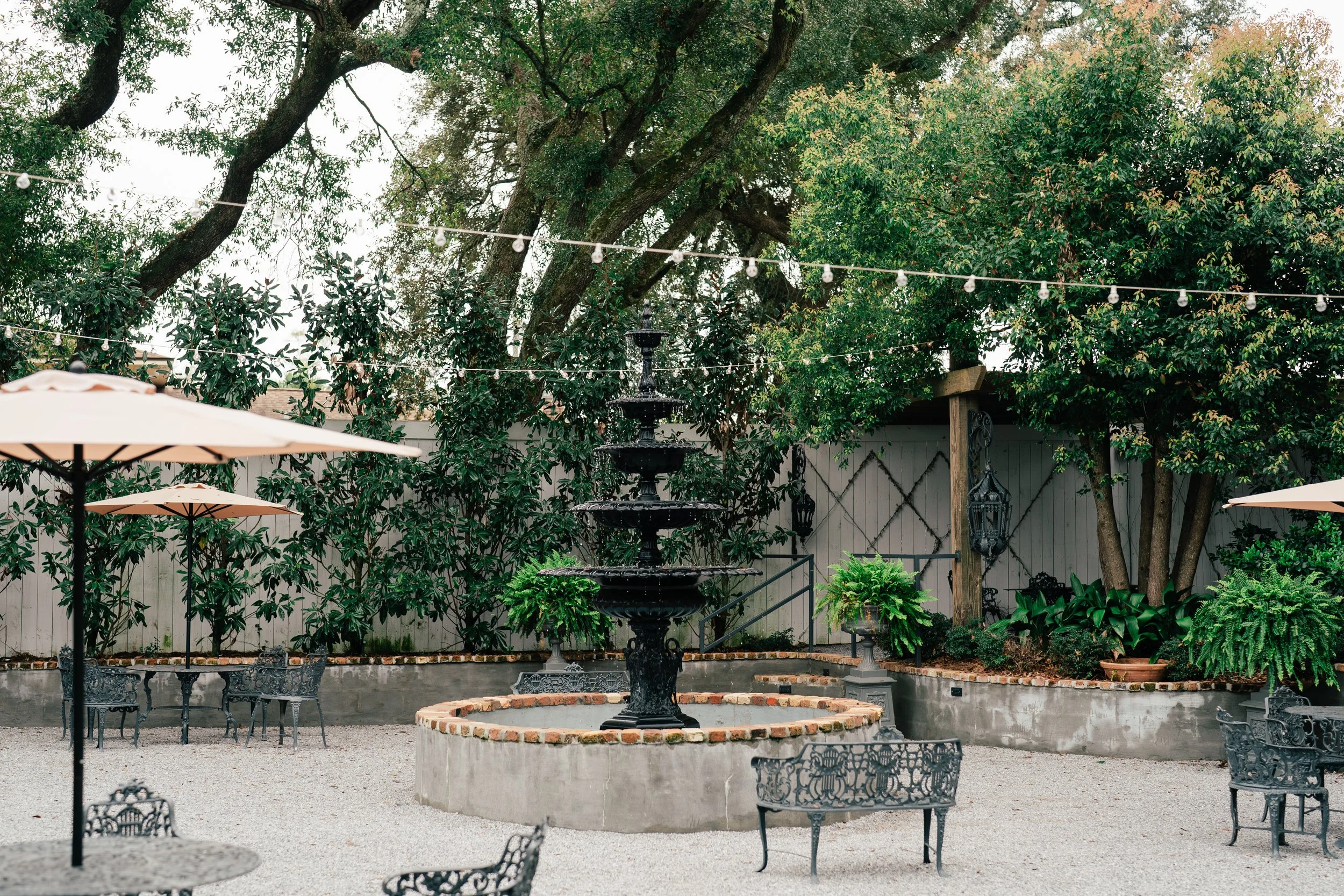 An outdoor garden area in a Pensacola Wedding Venue with a gravel ground, black metal chairs, beige umbrellas, a central black fountain, and surrounding green plants and trees, all enclosed by a white fence.