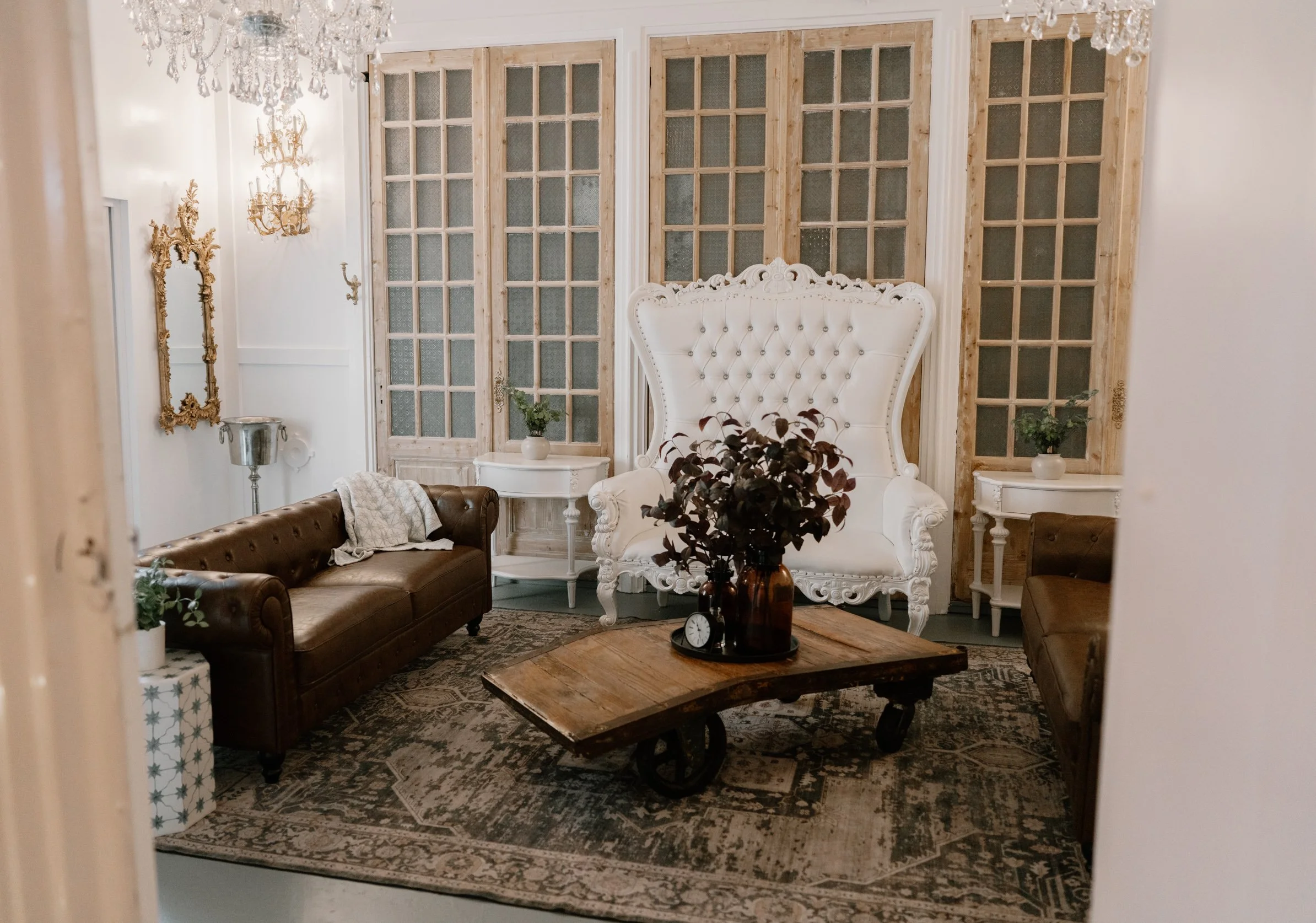 A cozy groom's suite featuring a high-backed white armchair with ornate detailing, two brown leather sofas, a rustic wooden coffee table with plant decorations, and vintage accents including a rug, gold-framed mirrors, and chandeliers.