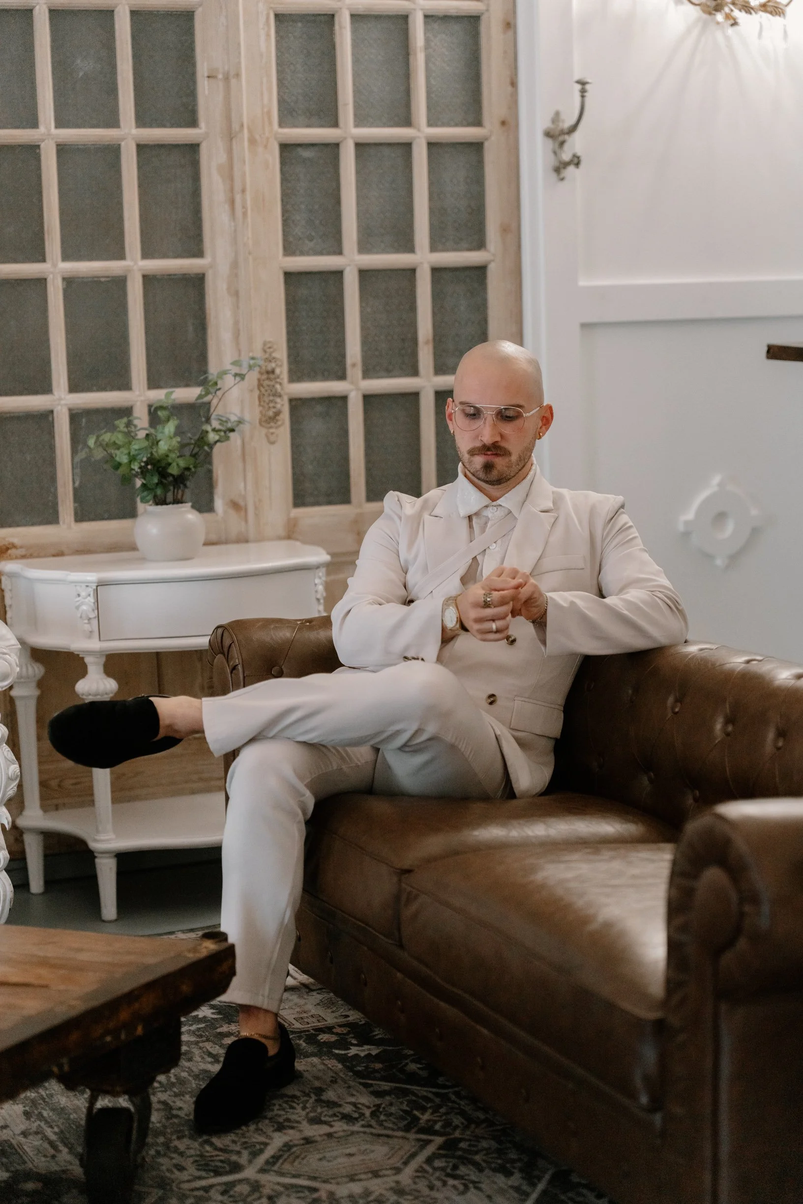 A man with glasses, a bald head, and a beard wearing a white suit, sitting on a brown leather sofa, looking at his watch, with one leg crossed over the other, in a vintage-style groom suite.