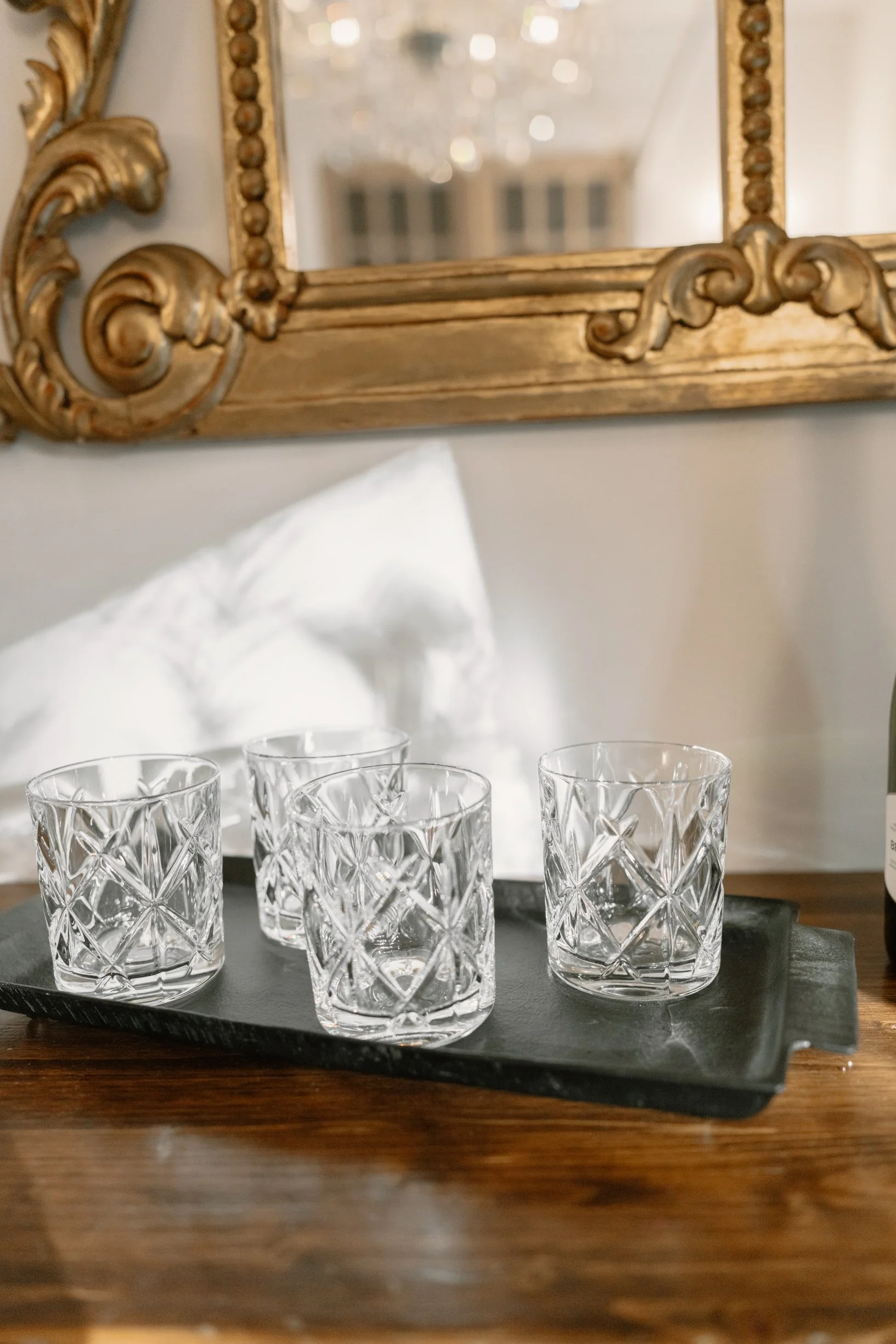Four cut-glass tumblers on a black tray, placed on a wooden surface with a white pillow and a mirror with a gold ornate frame in the background in a Pensacola Wedding Venue.