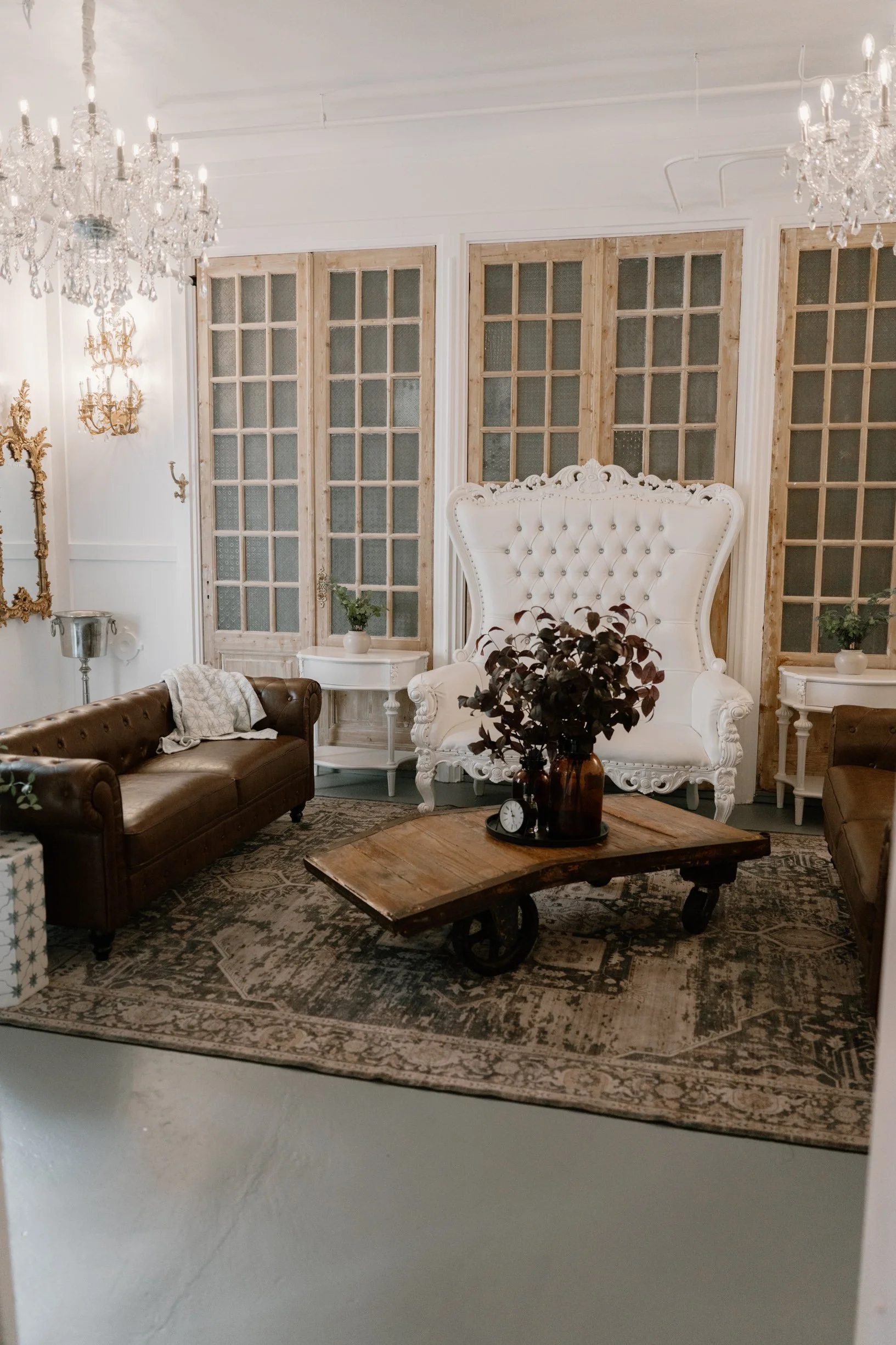 Elegant groom suite with a large white tufted armchair, brown leather sofas, vintage wooden coffee table with a vase of dark leaves, ornate gold mirror, chandeliers, and decorative screens.