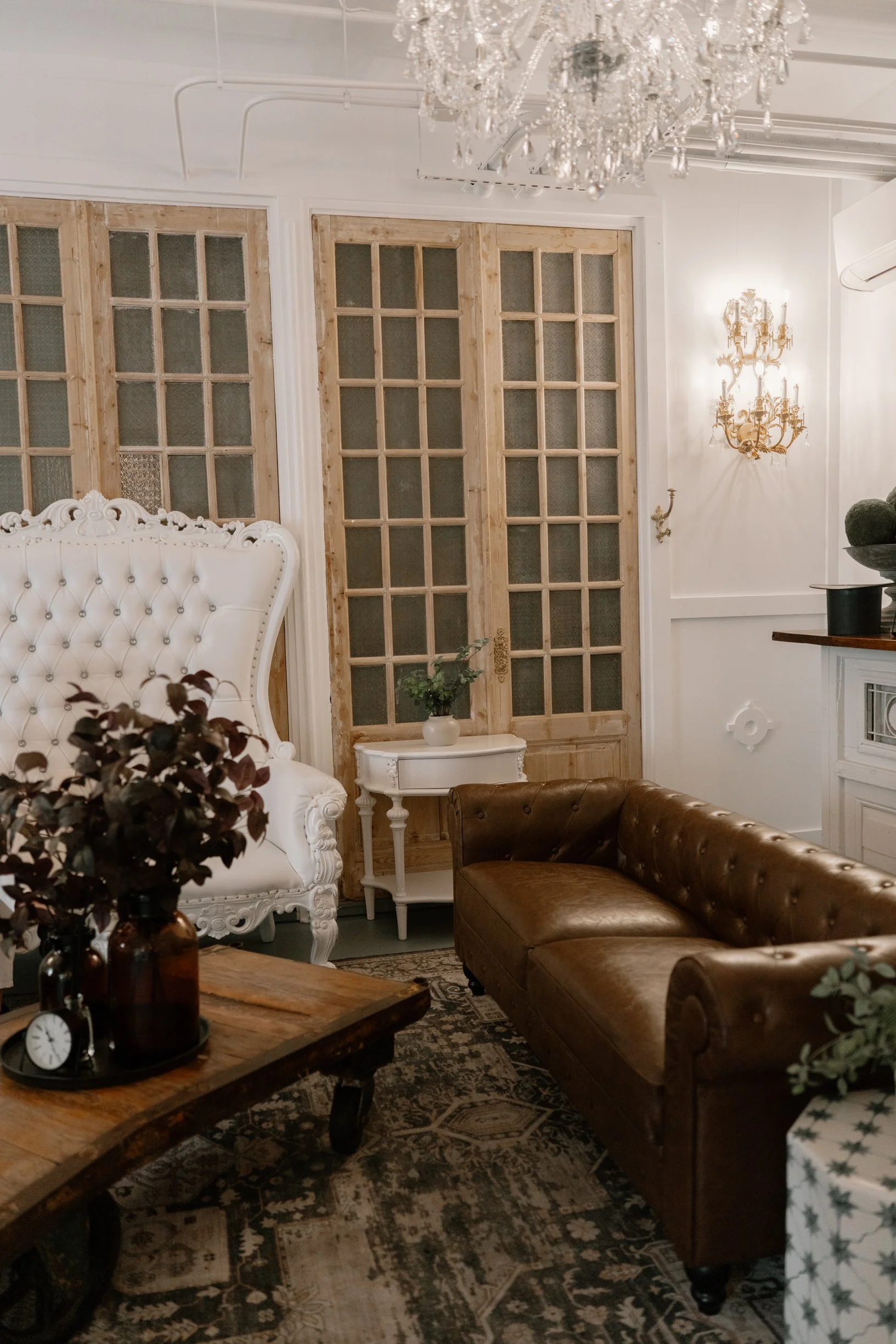 A living room corner with a vintage tufted white armchair, a brown leather sofa, a wooden coffee table with a vase of dried flowers and a clock, a white side table with a small potted plant, features wooden sliding doors, a chandelier, wall sconces, 