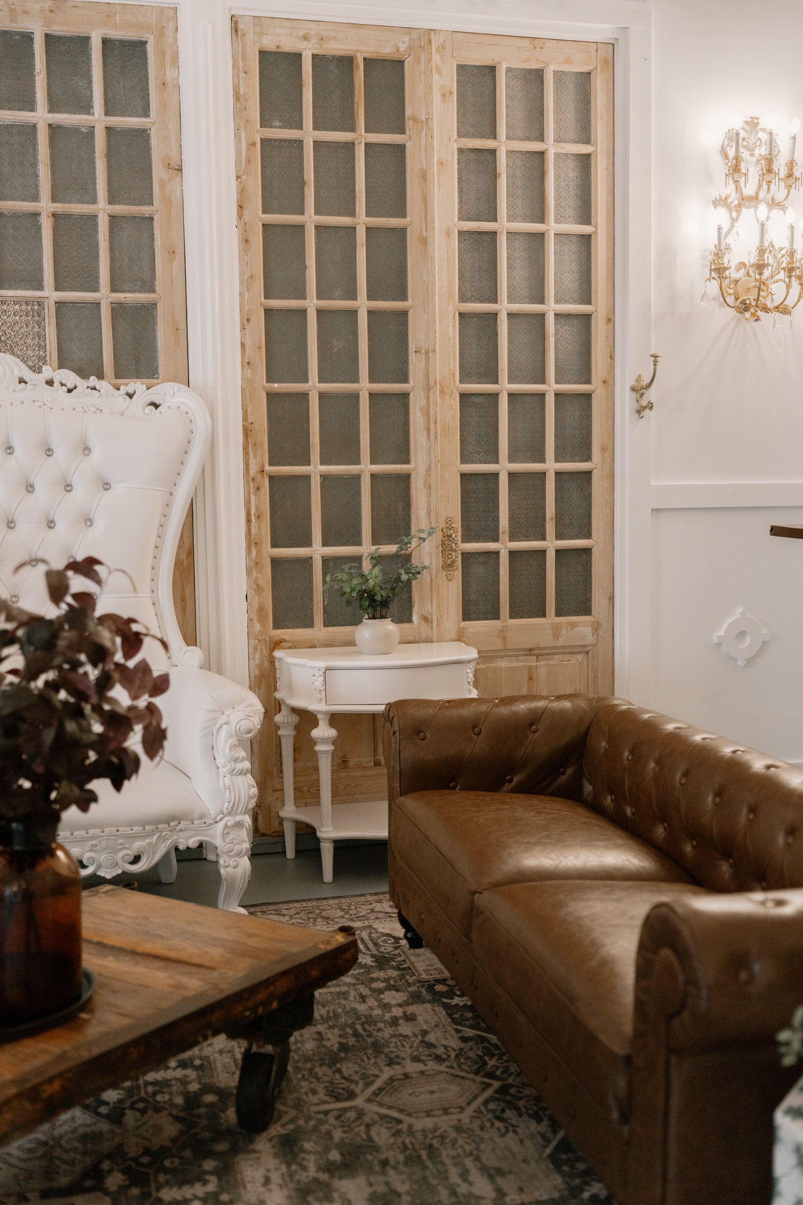 A vintage living room corner with a white ornate high-back chair, a brown leather tufted sofa, a rustic wooden coffee table with iron wheels, a white side table with a potted plant, and decorative wooden sliding doors with glass panes. There is a gol