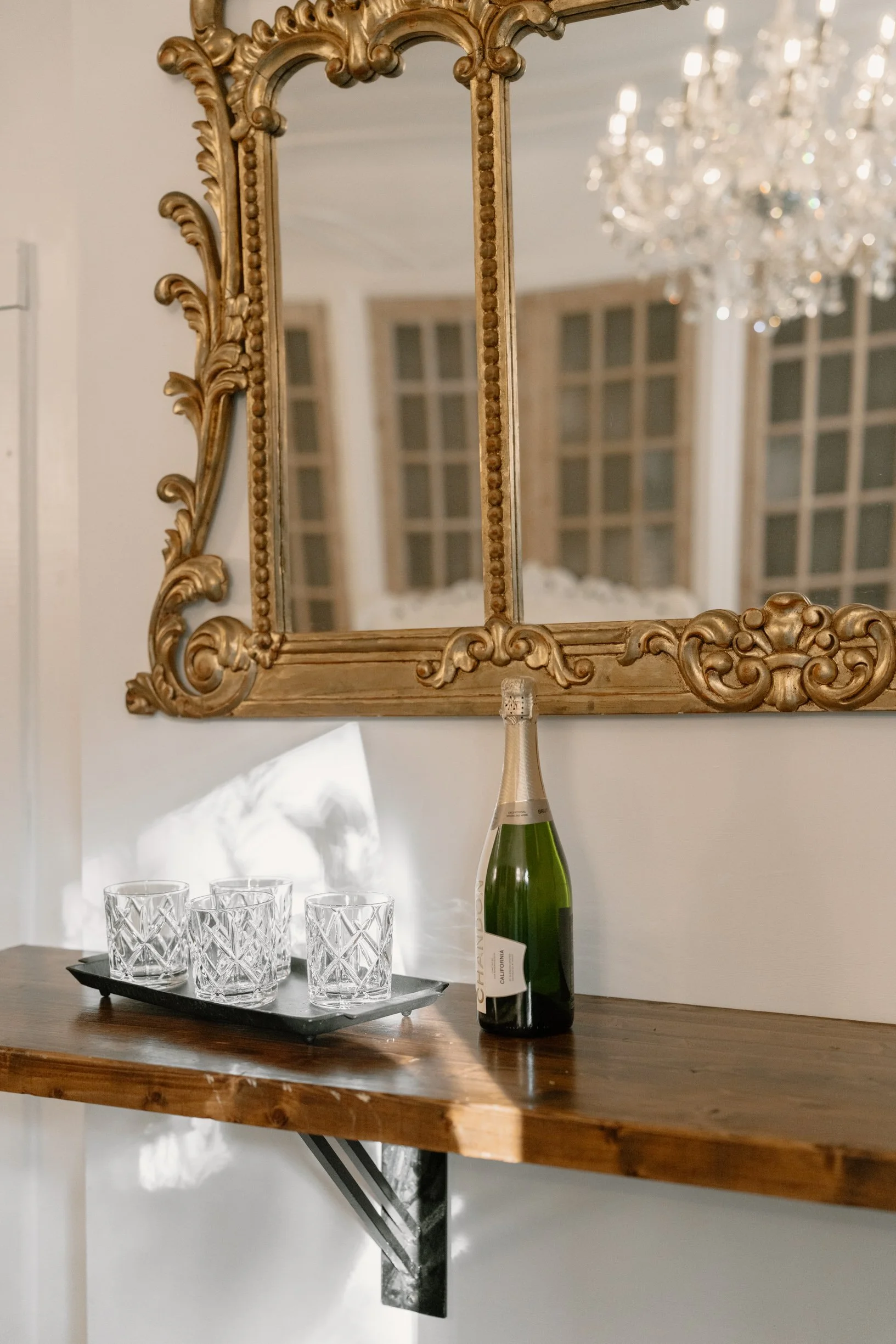 A wooden console table with four crystal glasses on a tray and a bottle of champagne. A large, ornate gold-framed mirror hangs above the table, reflecting a room with a chandelier and windows in a Pensacola Wedding Venue.