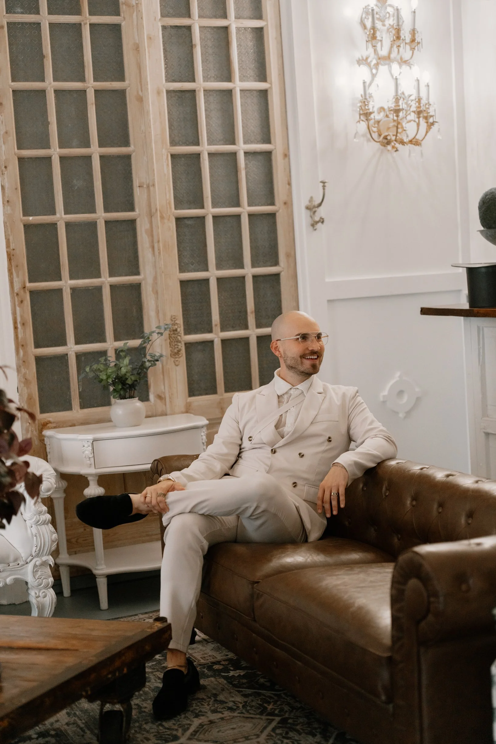 A man dressed in an elegant white suit sitting on a brown leather sofa, smiling and looking to his left in a vintage-style interior with ornate wall details and a chandelier in a Pensacola Wedding Venue.