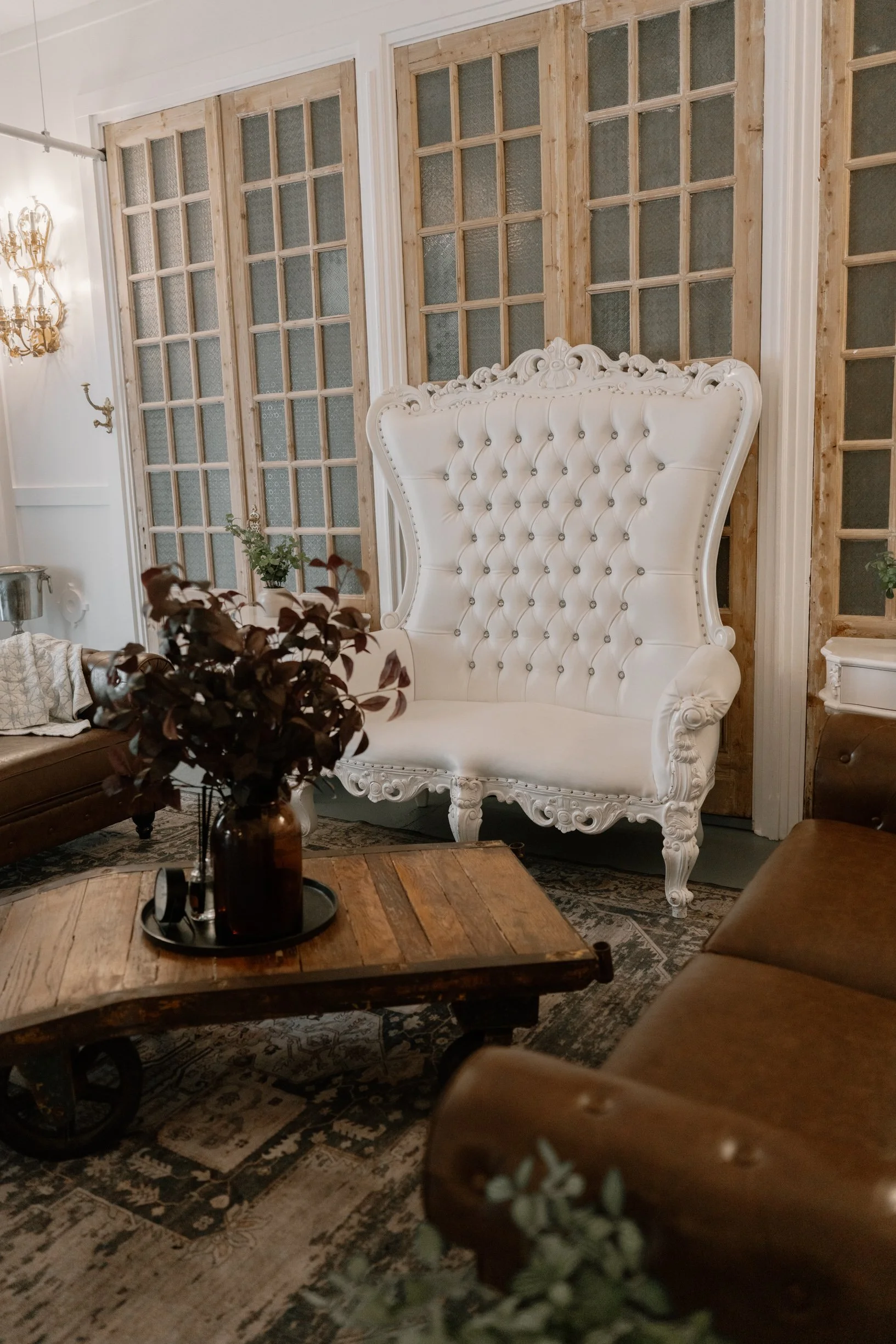 A vintage-style groom suite with a white tufted high-back chair, wooden sliding doors, a wooden coffee table, and a dark brown sofa in a Pensacola Wedding Venue.