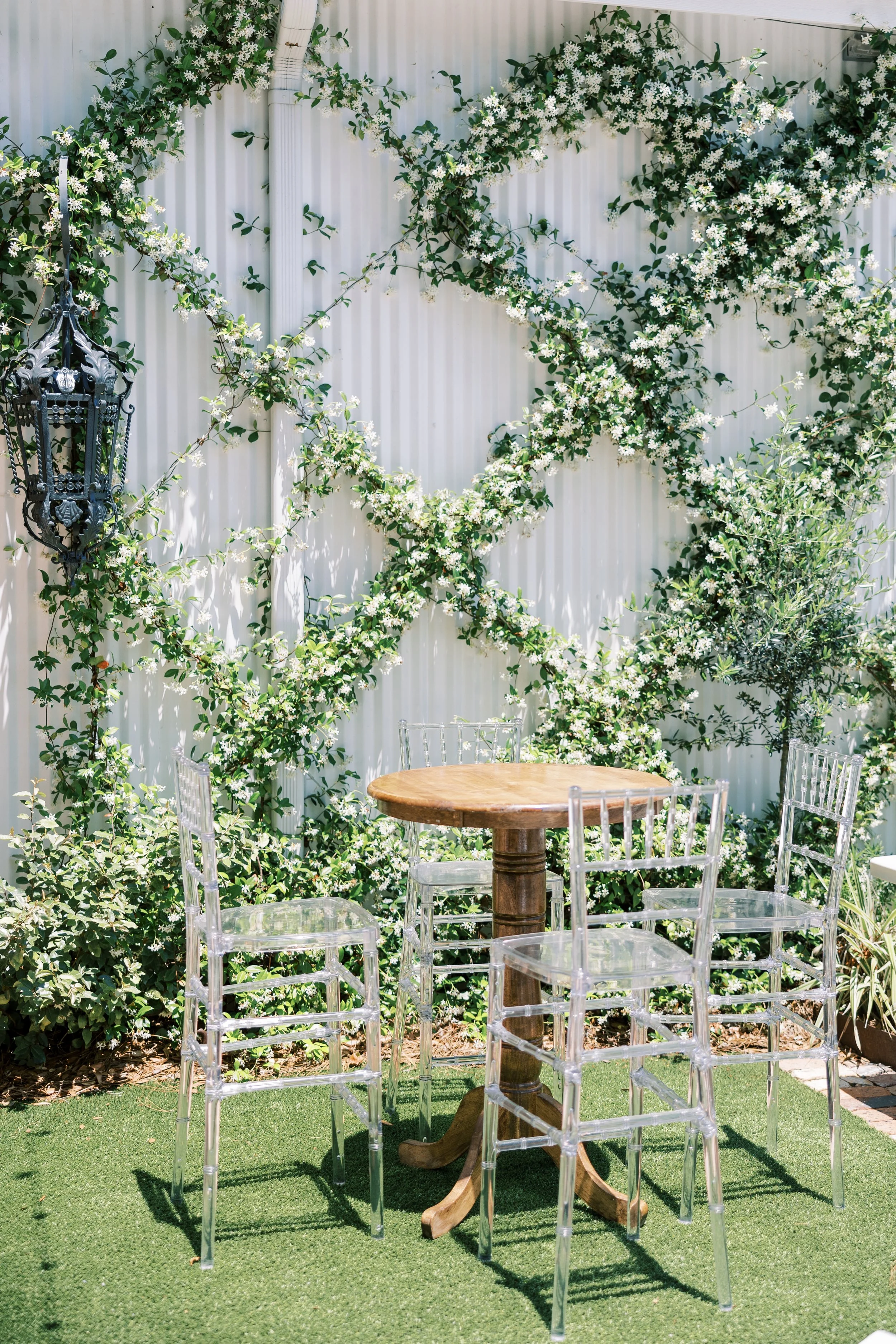 Round wooden table with transparent chairs in a garden with white flowering vines and greenery on a white fence.