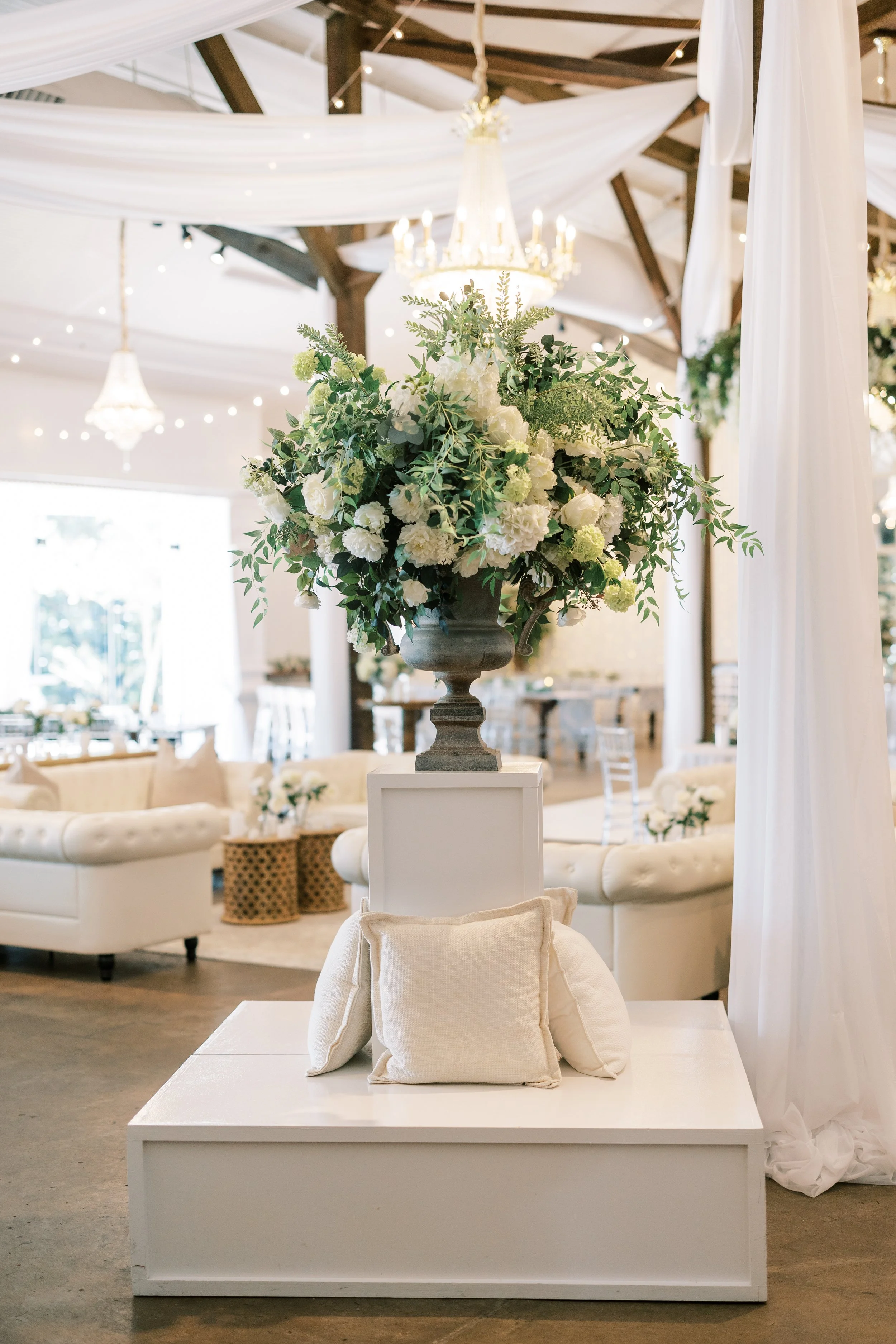 Elegant wedding or event decor featuring a large floral arrangement with white flowers and green foliage on a pedestal, set on a white platform with pillows, in a bright, decorated indoor space with chandeliers and draped fabric.