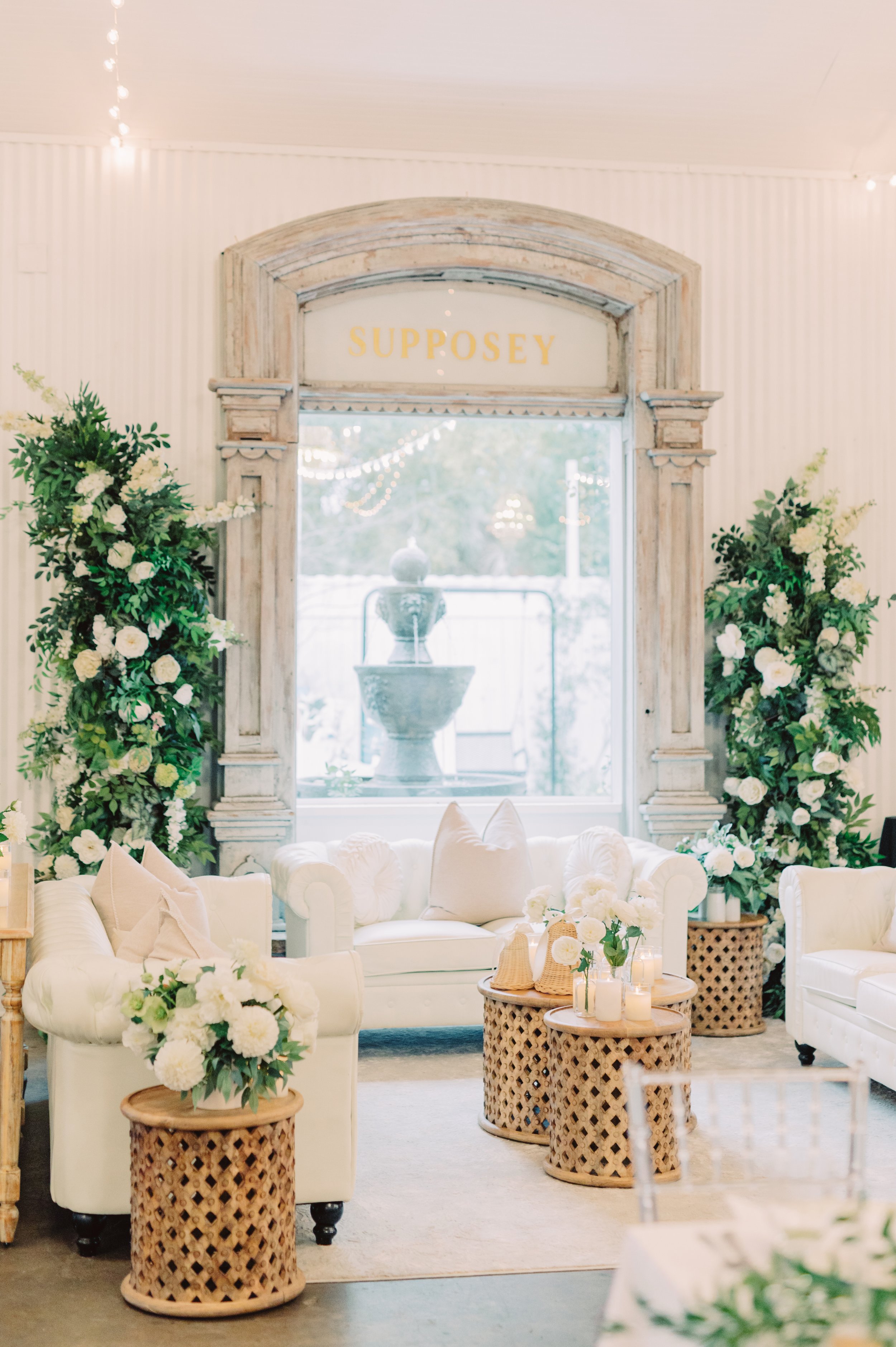 Elegant indoor event space with white sofas, floral arrangements, and natural wicker side tables, featuring a large window with greenery and a fountain outside.