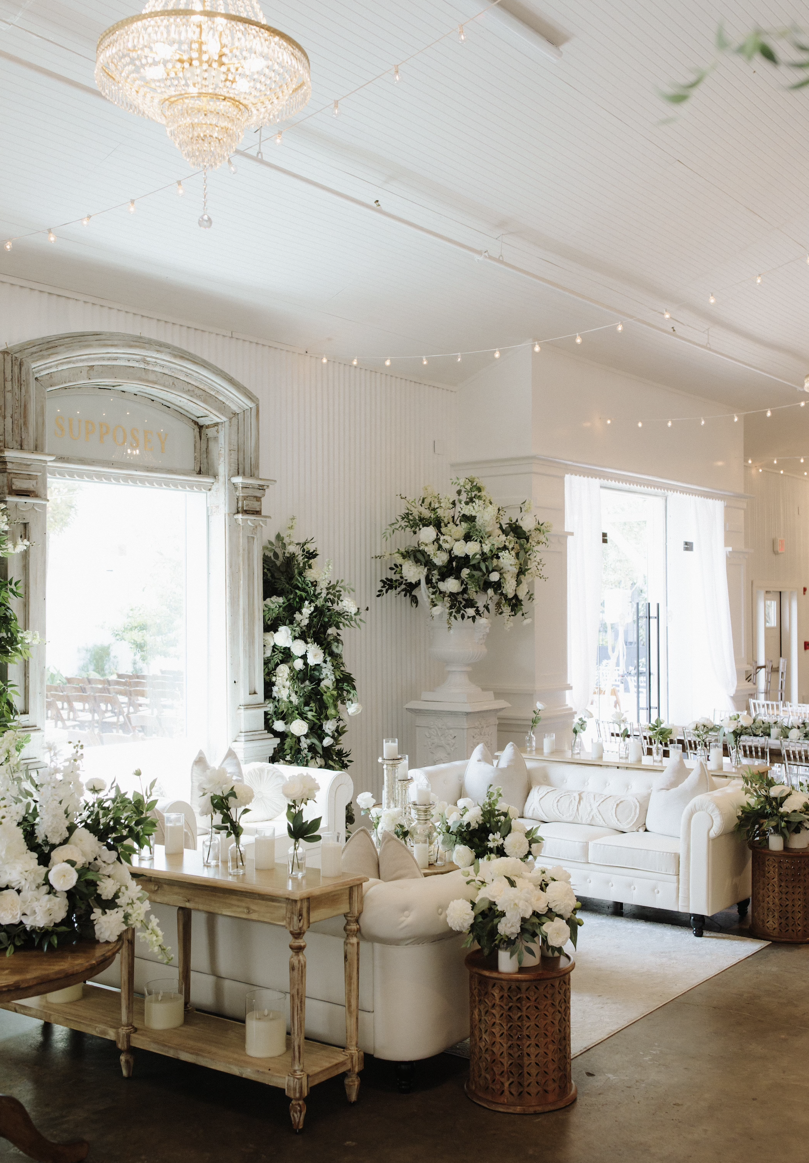 Elegant event room decorated in white with floral arrangements, candles, and vintage furniture, featuring a large chic chandelier and string lights overhead.