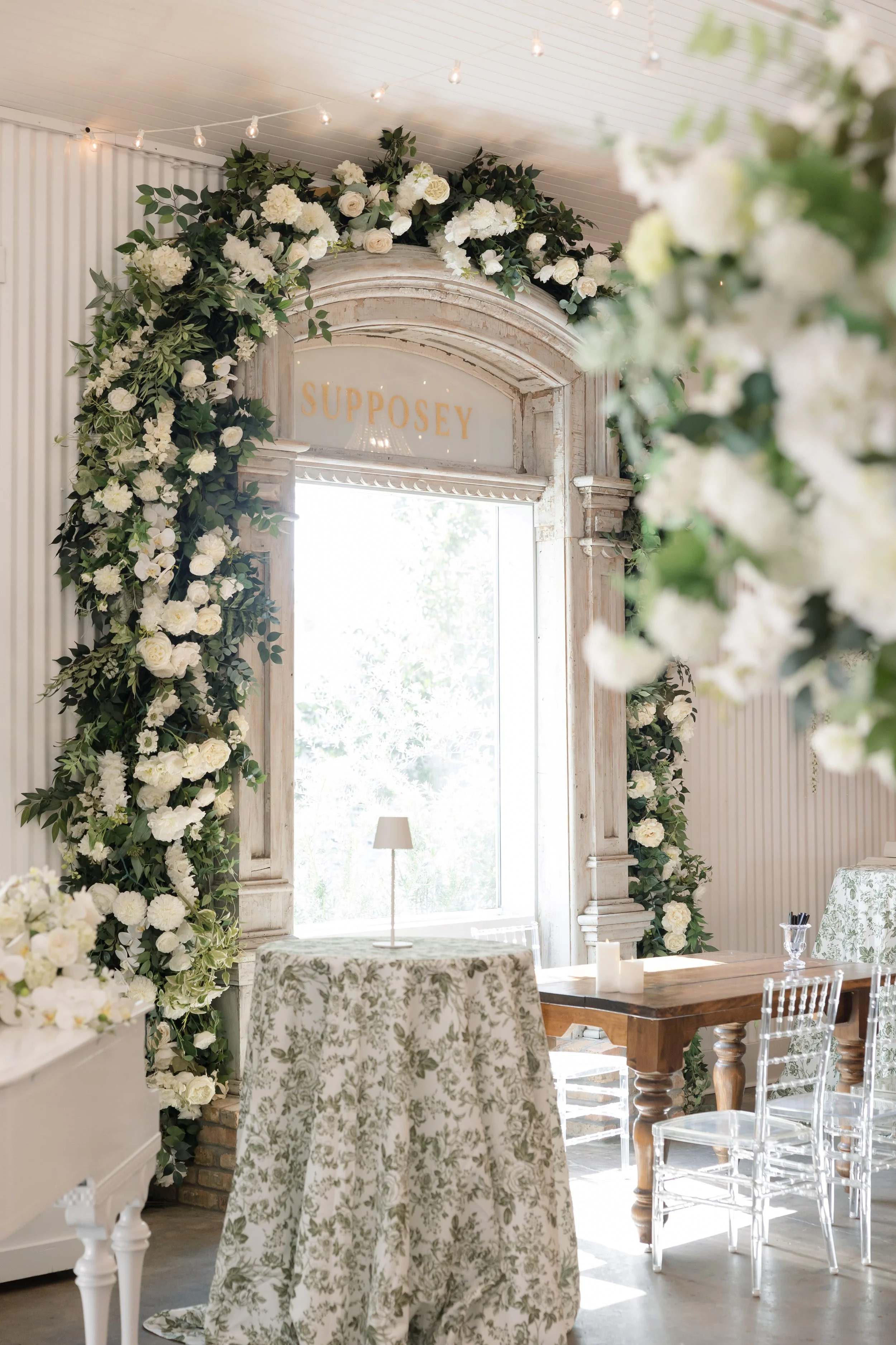 Elegant indoor space decorated with white and green floral arrangements around a window frame with a sign that reads "SUPOSEY" above it. The room features tables draped with patterned tablecloths, illuminated by natural light and string lights on the