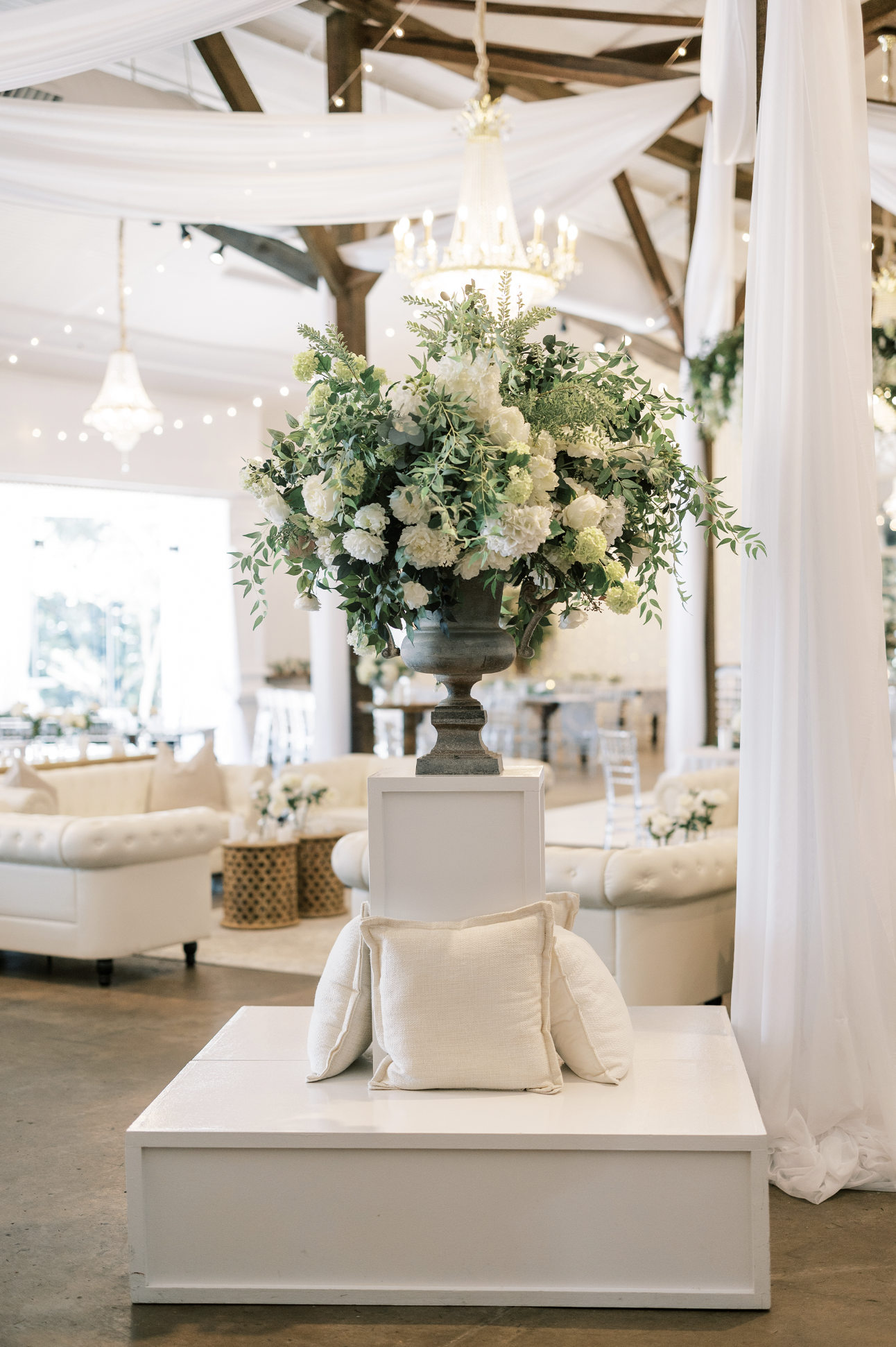 Elegant wedding or event decor featuring a large floral arrangement on a gray pedestal, surrounded by white cushioned furniture and soft lighting in a bright, airy indoor Pensacola Wedding Venue.