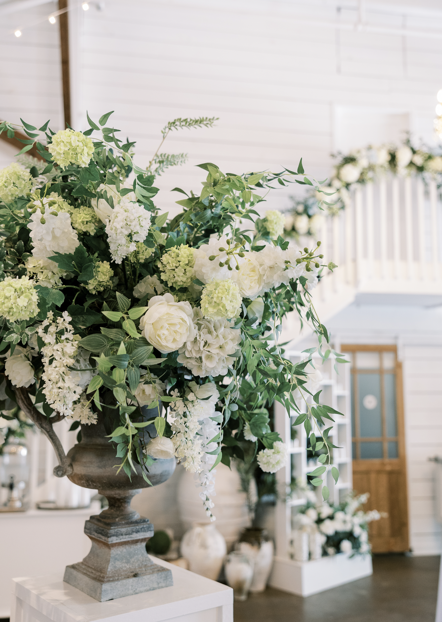 Large white floral arrangement with roses, hydrangeas, and greenery in a stone urn on a white pedestal inside a bright, white-paneled Pensacola Wedding Venue.