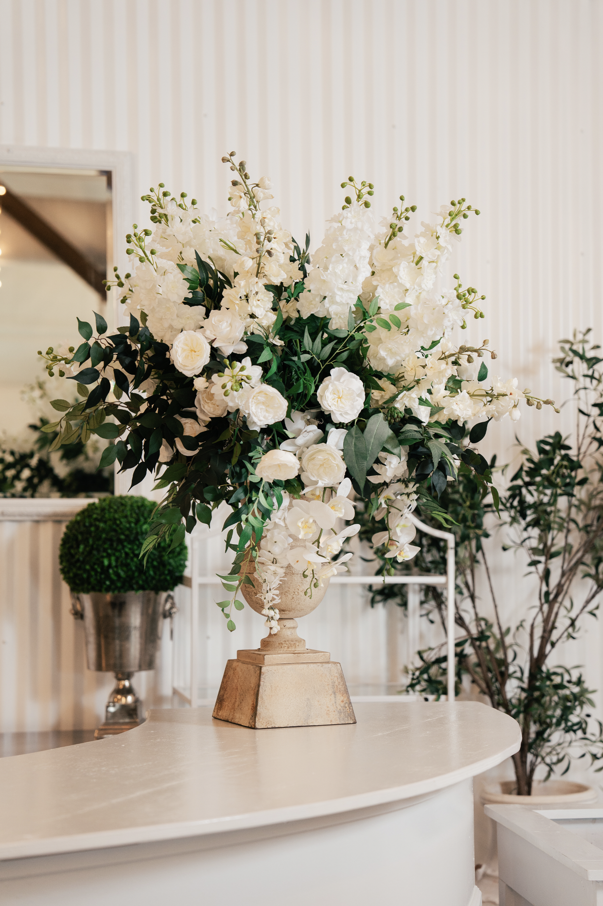 A large white floral arrangement with roses and orchids in a decorative beige vase on a white curved table. A green potted plant is visible in the background against a beige striped wall in a Pensacola Wedding Venue.