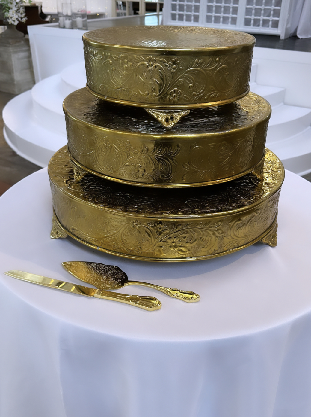 Three stacked ornate gold cake tins with floral patterns, on a white tablecloth, accompanied by a gold cake server and knife.