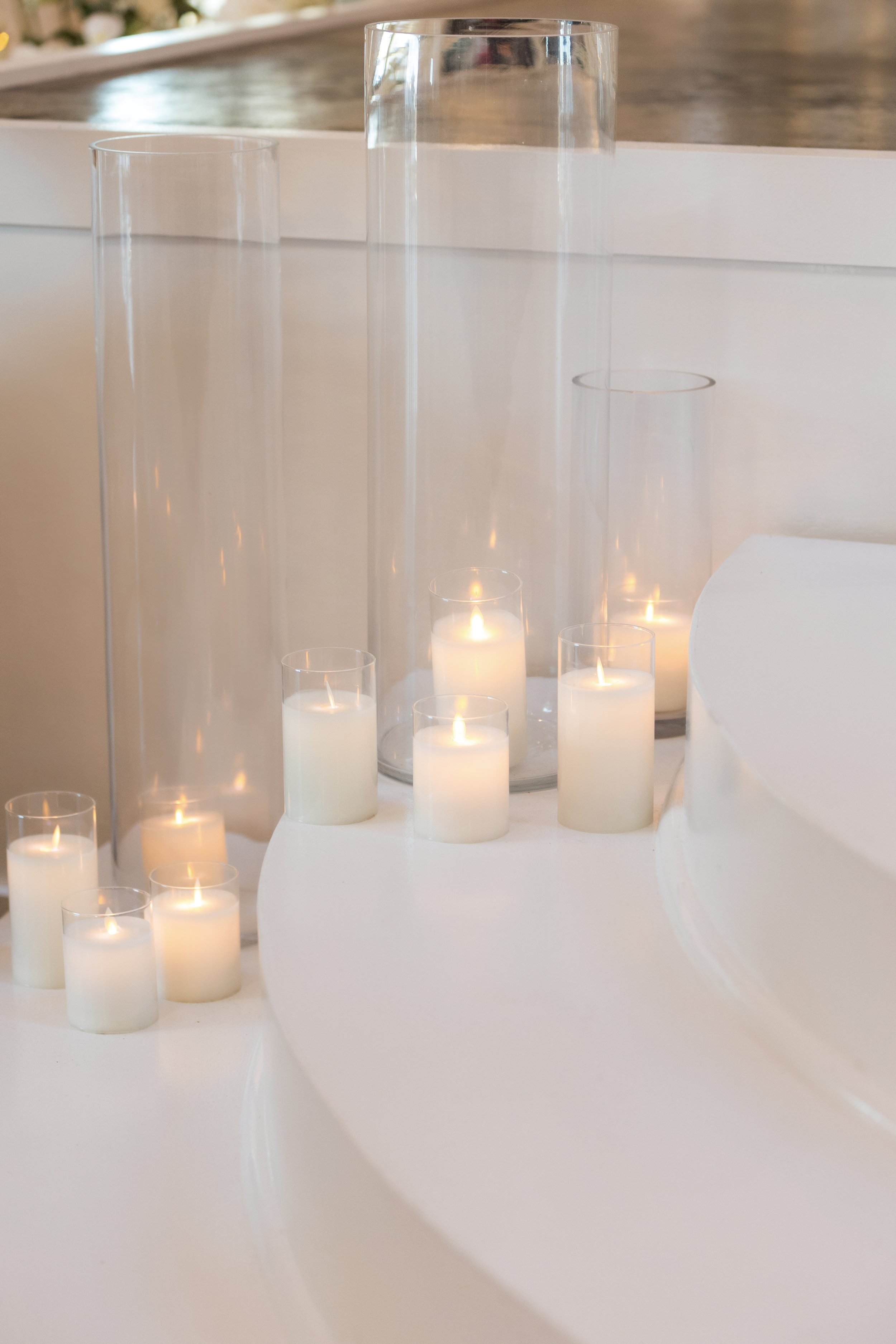 Decorative scene with lit white candles and empty glass vases of varying heights on a white surface.