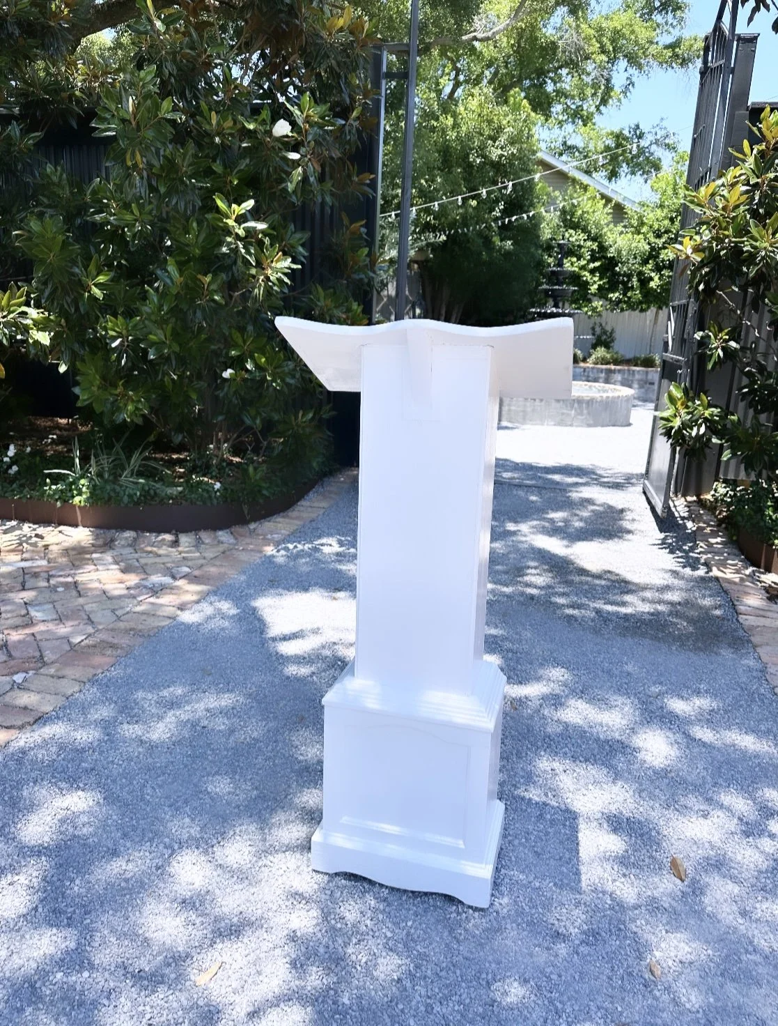 A white podium stands outdoors on a gravel surface, with a garden and trees in the background, and a string of lights overhead.