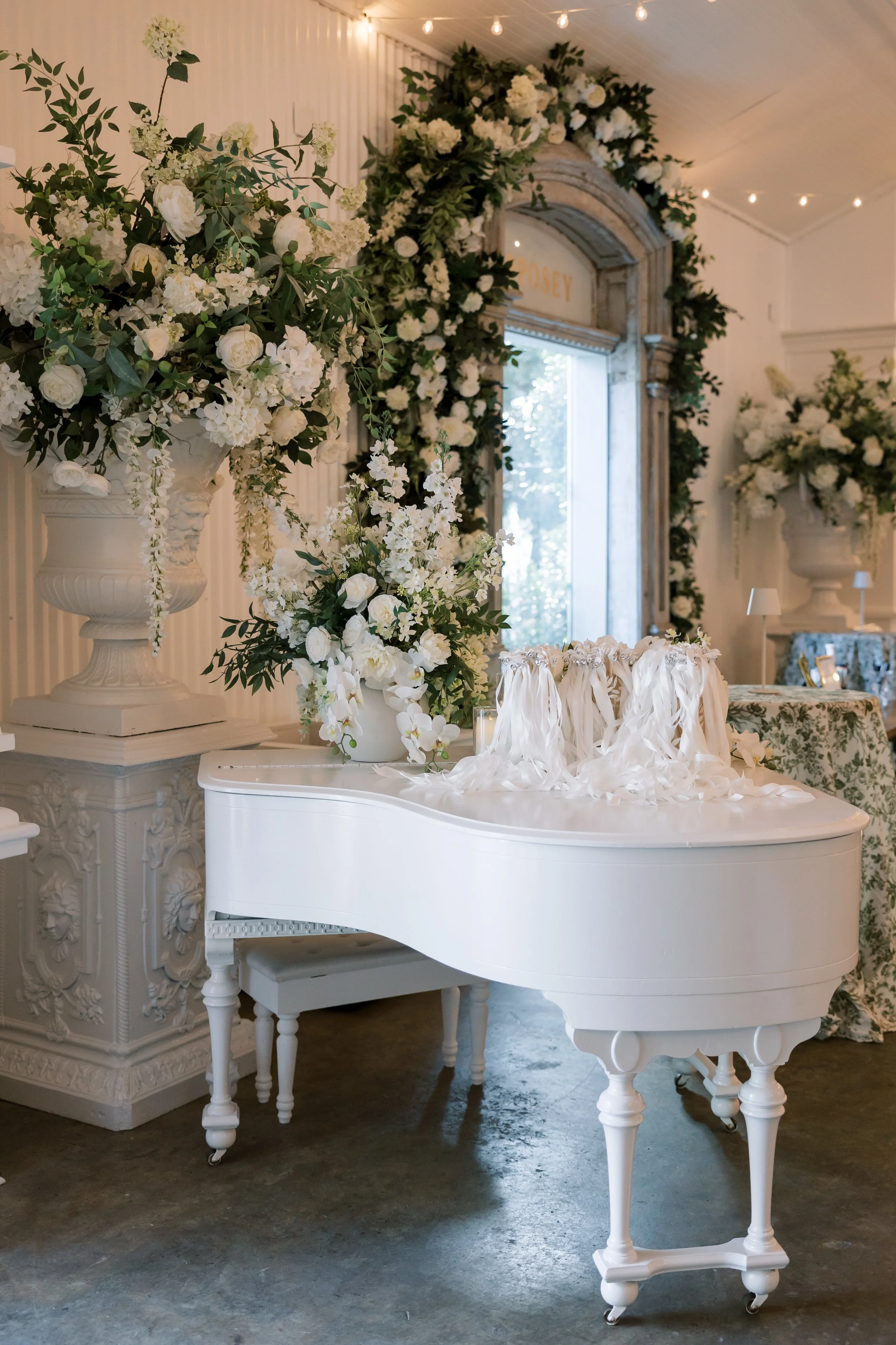 White grand piano with a floral arrangement on top, in a decorated room with large vases filled with white flowers and greenery, and a large mirror with floral decorations in the background of a Pensacola Wedding Venue.