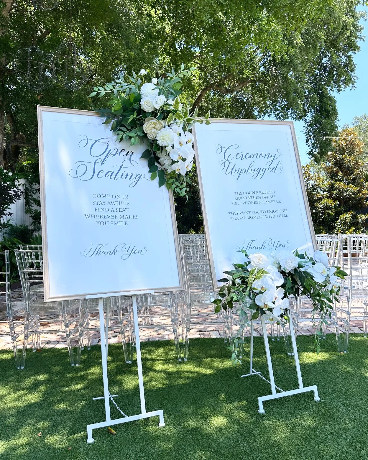 Pensacola Wedding ceremony signage with floral decorations on a sunny outdoor lawn, featuring signs labeled 'Open Seating' and 'Ceremony Unplugged' with chairs arranged in the background.