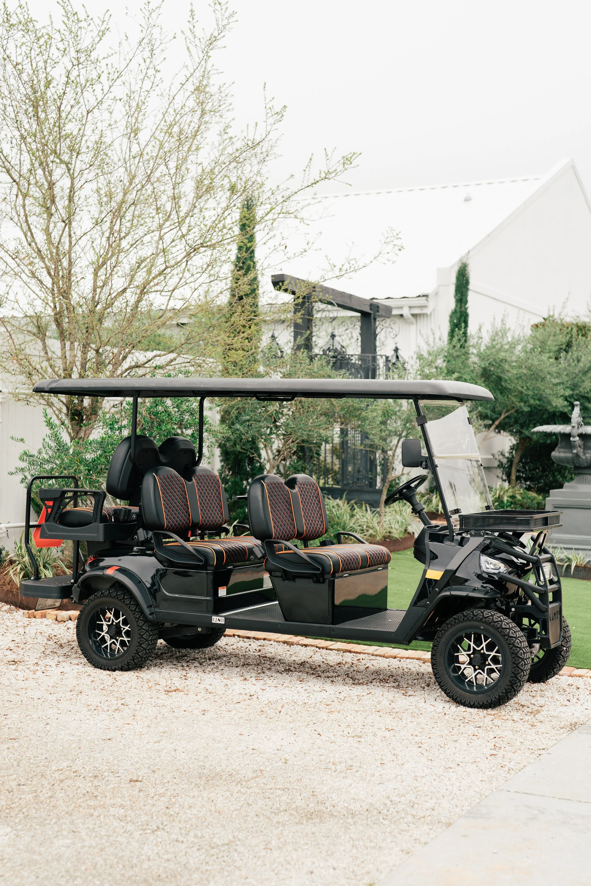 A black golf cart with four seats, parked on a gravel surface, with a house, trees, and a fountain in the background.